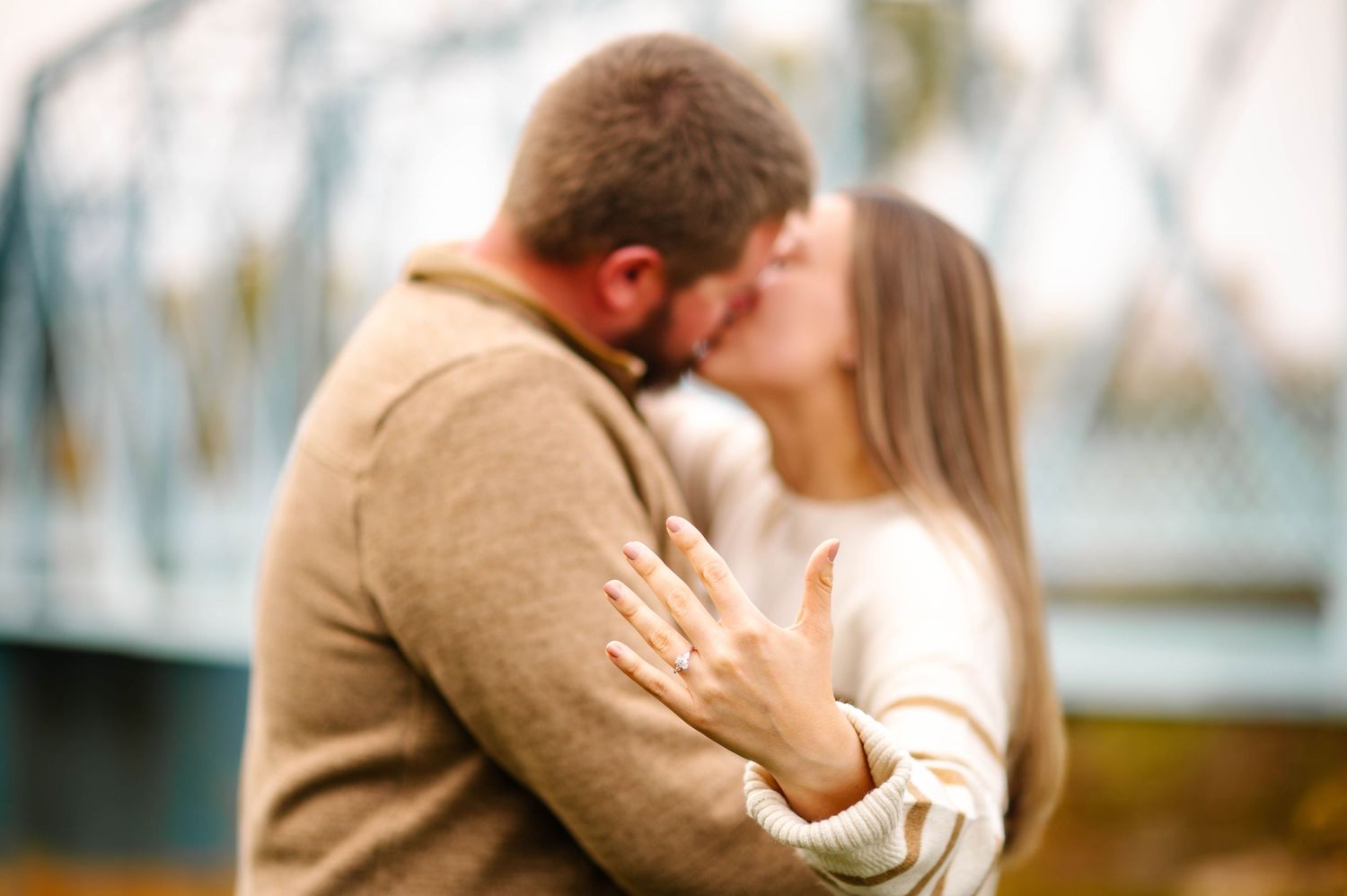 A couple embraces during an autumn engagement photoshoot near a bridge with fall foliage in the background.