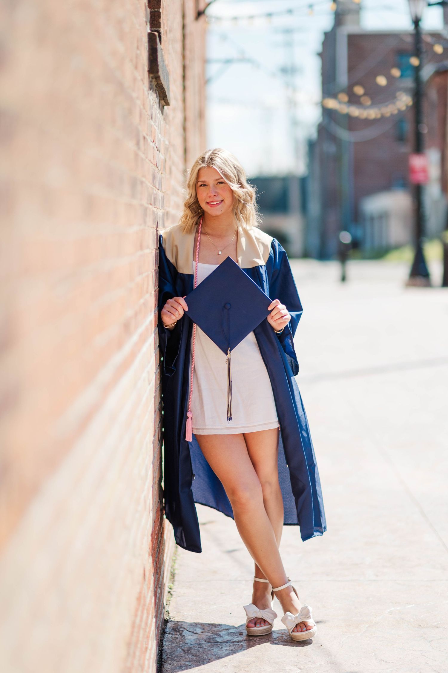 Smiling blonde graduate in navy cap and gown holding mortarboard against brick wall outdoors.