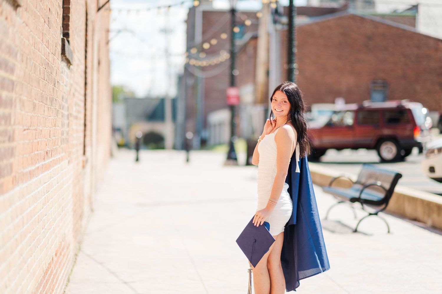 Young woman in white dress carrying navy graduation gown on sunny brick alley street.
