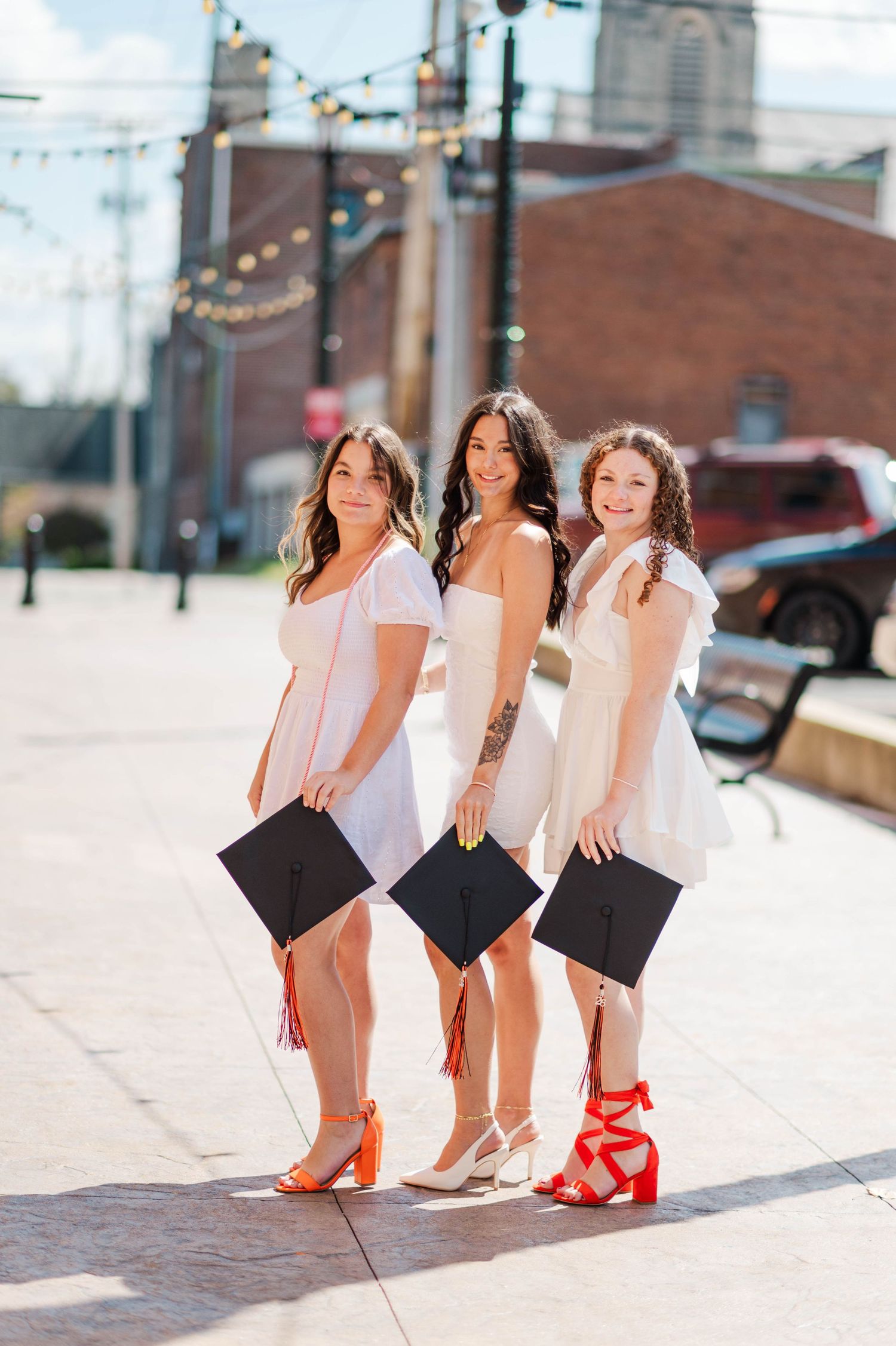 Three smiling graduates in white dresses hold black caps outdoors on a sunny brick plaza.