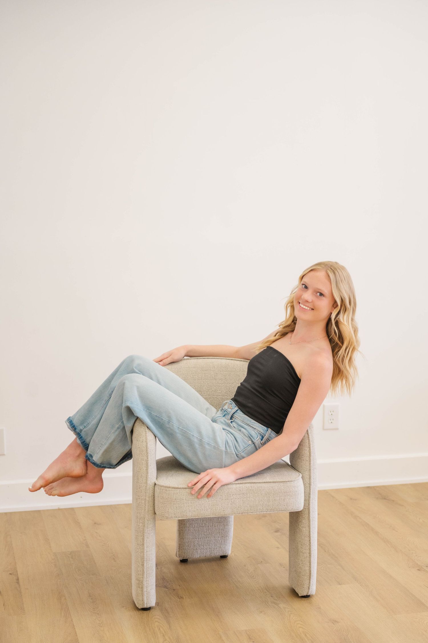 Blonde woman in black tube top and light wash jeans sitting relaxed in a modern chair.
