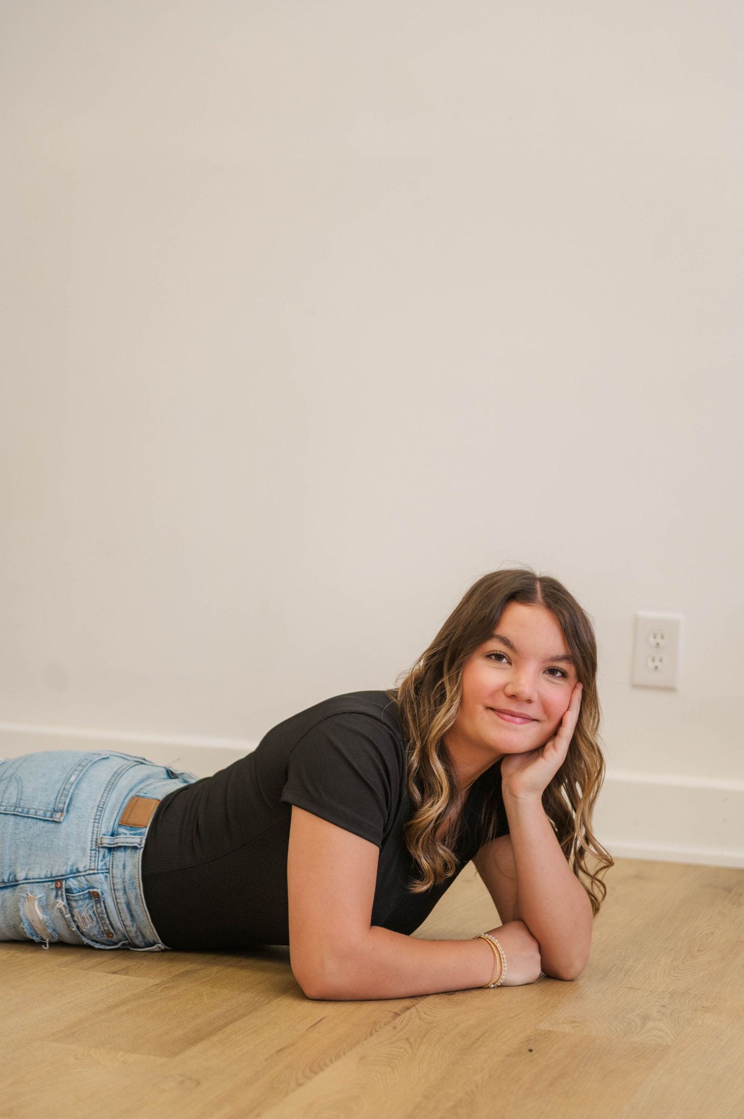 Smiling young woman in black tee and ripped jeans lying on hardwood floor, resting chin on hand.