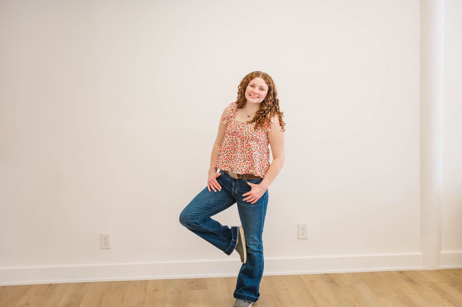 Curly-haired woman in floral top and dark jeans posing with one leg raised, smiling.