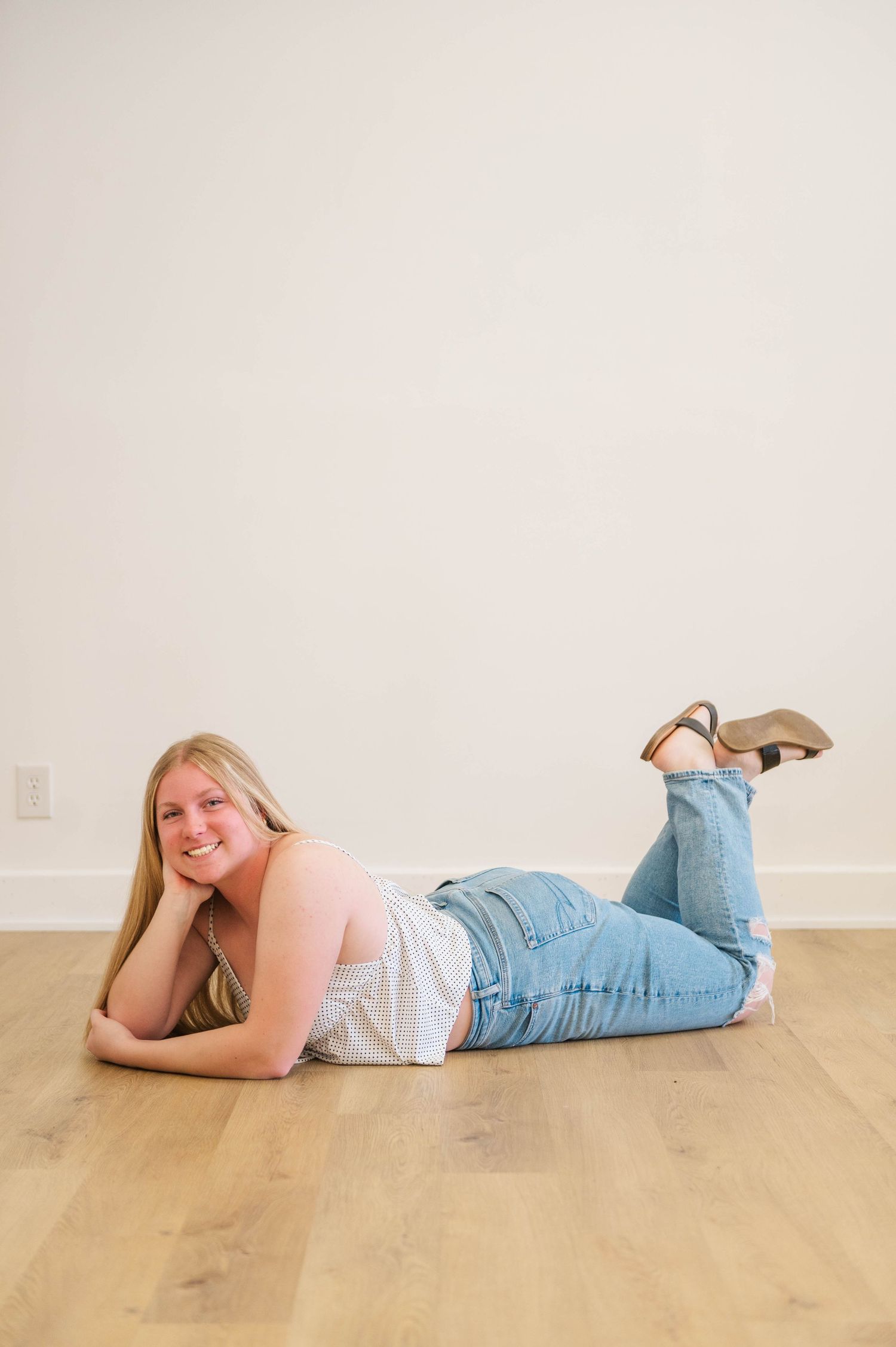 Smiling blonde woman lying on hardwood floor in white top and blue jeans with heels.