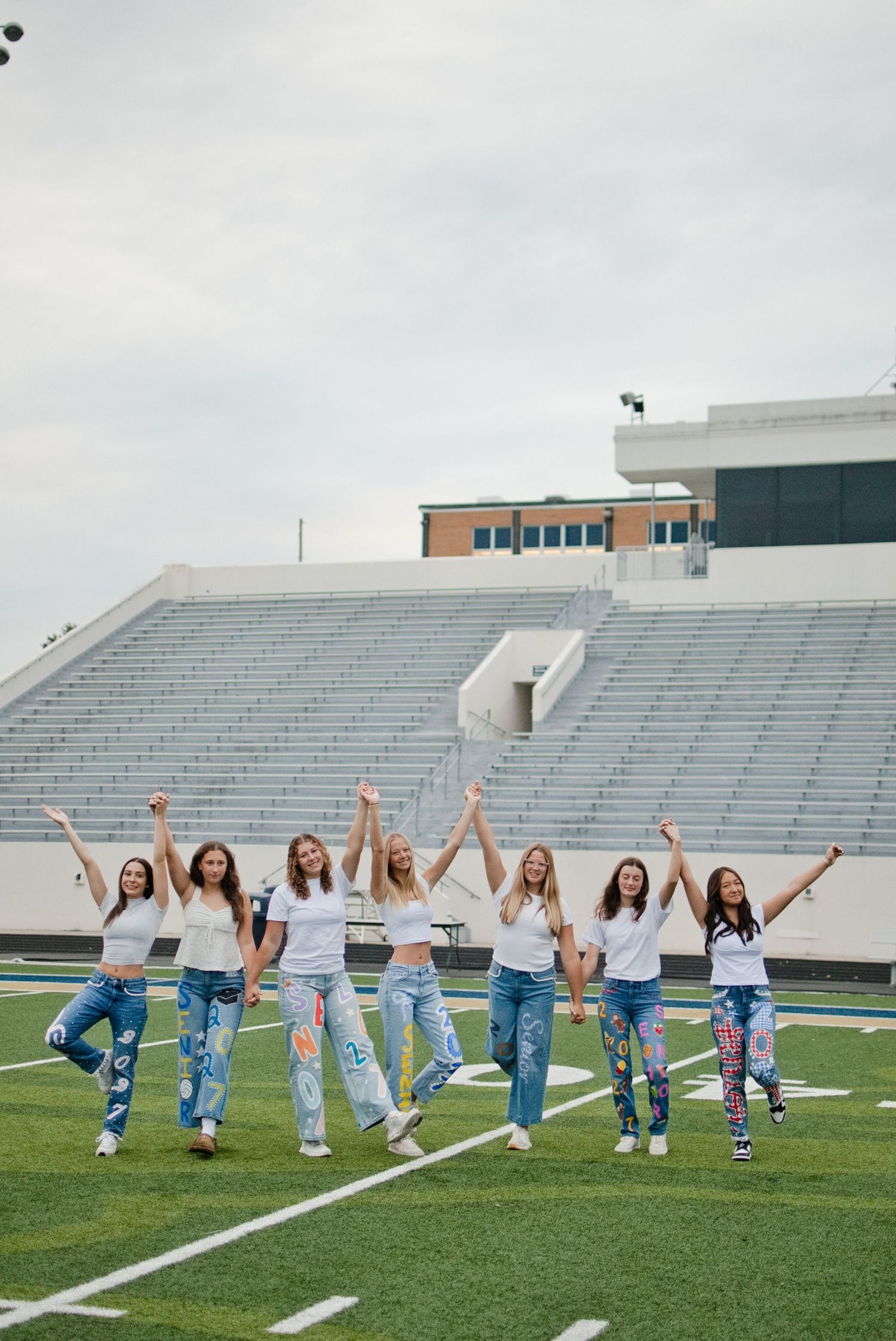 Eight young women in white tees and painted jeans celebrate with raised arms on a football field stadium.