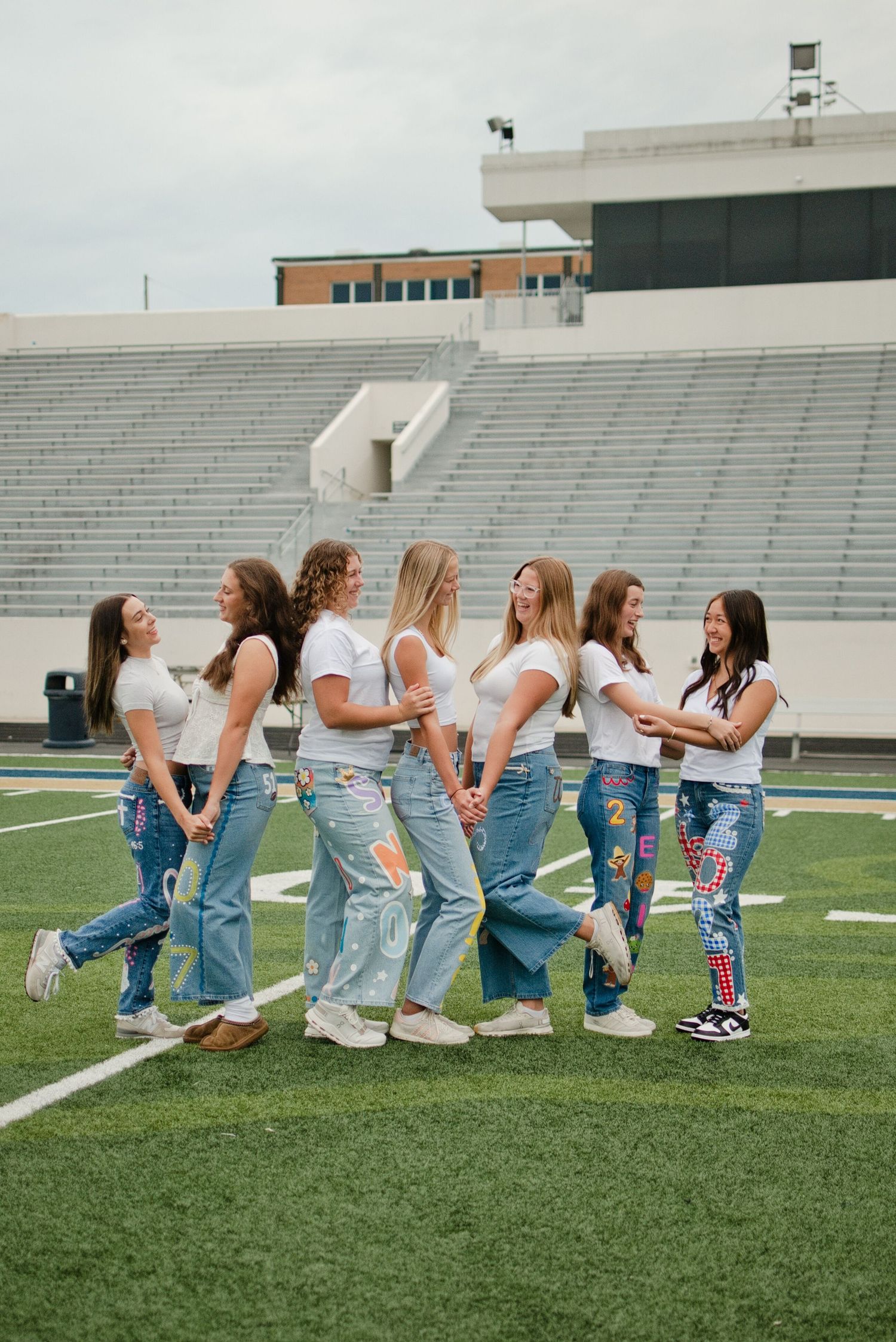 Seven teenage girls in white tees and decorated jeans hold hands on a football field stadium.