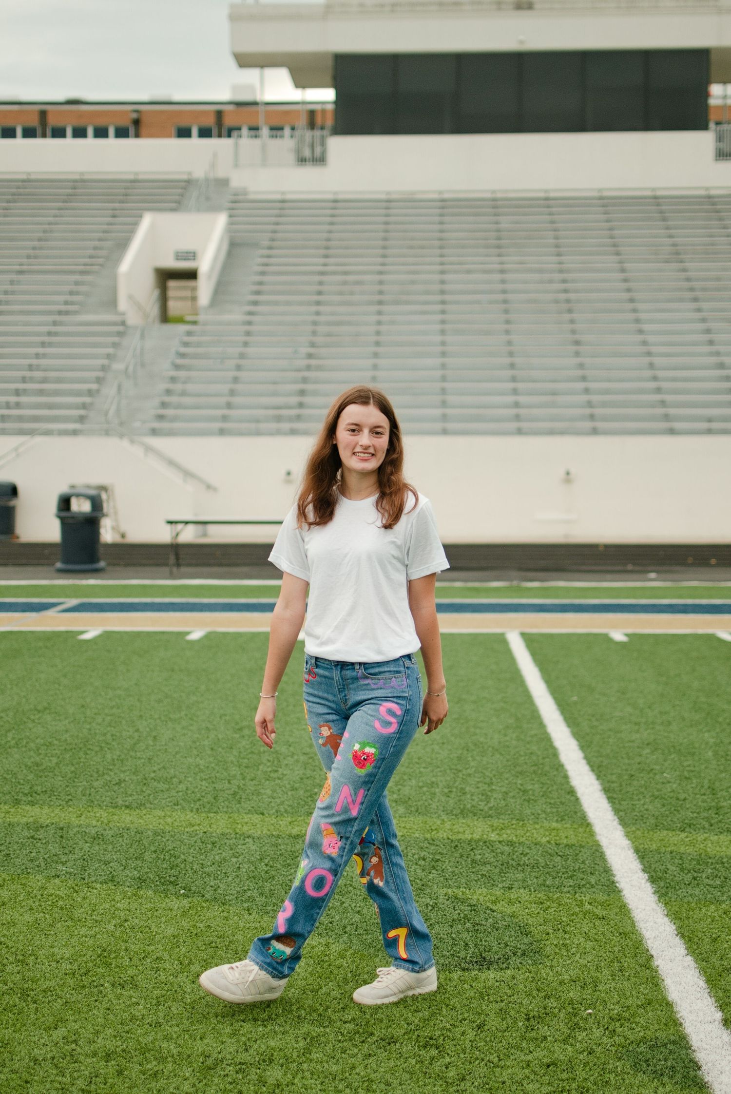 Teen girl in white t-shirt and colorful custom senior jeans smiling while walking on a football field.