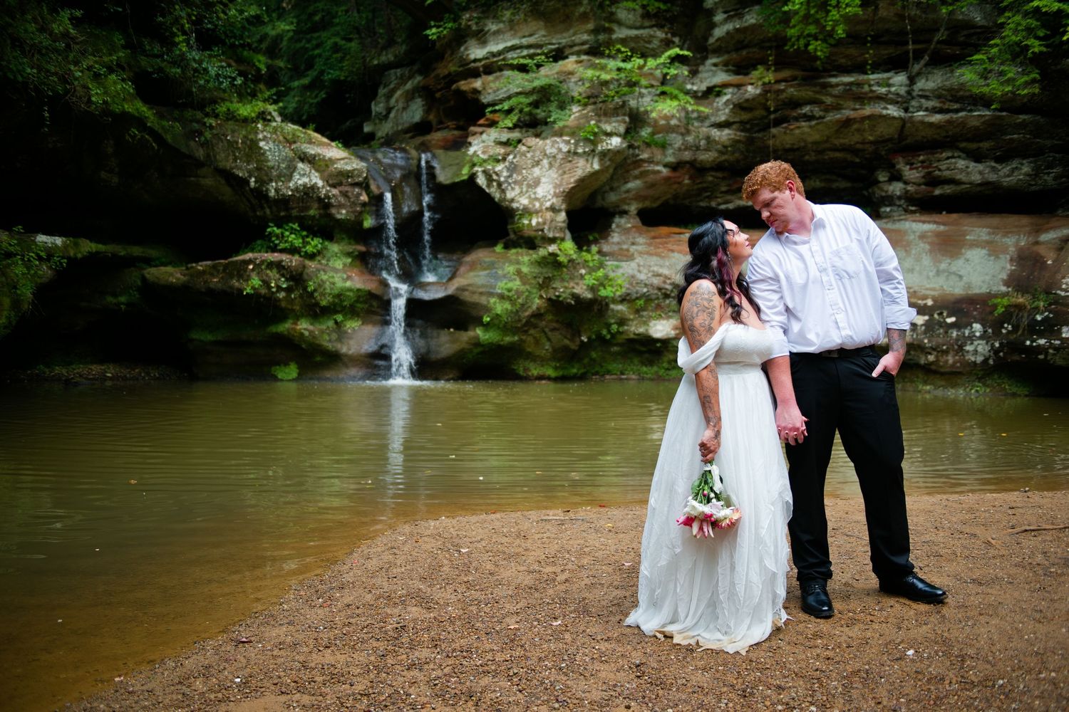 Bride and groom share a romantic moment by a waterfall in a lush green forest setting on their wedding day.