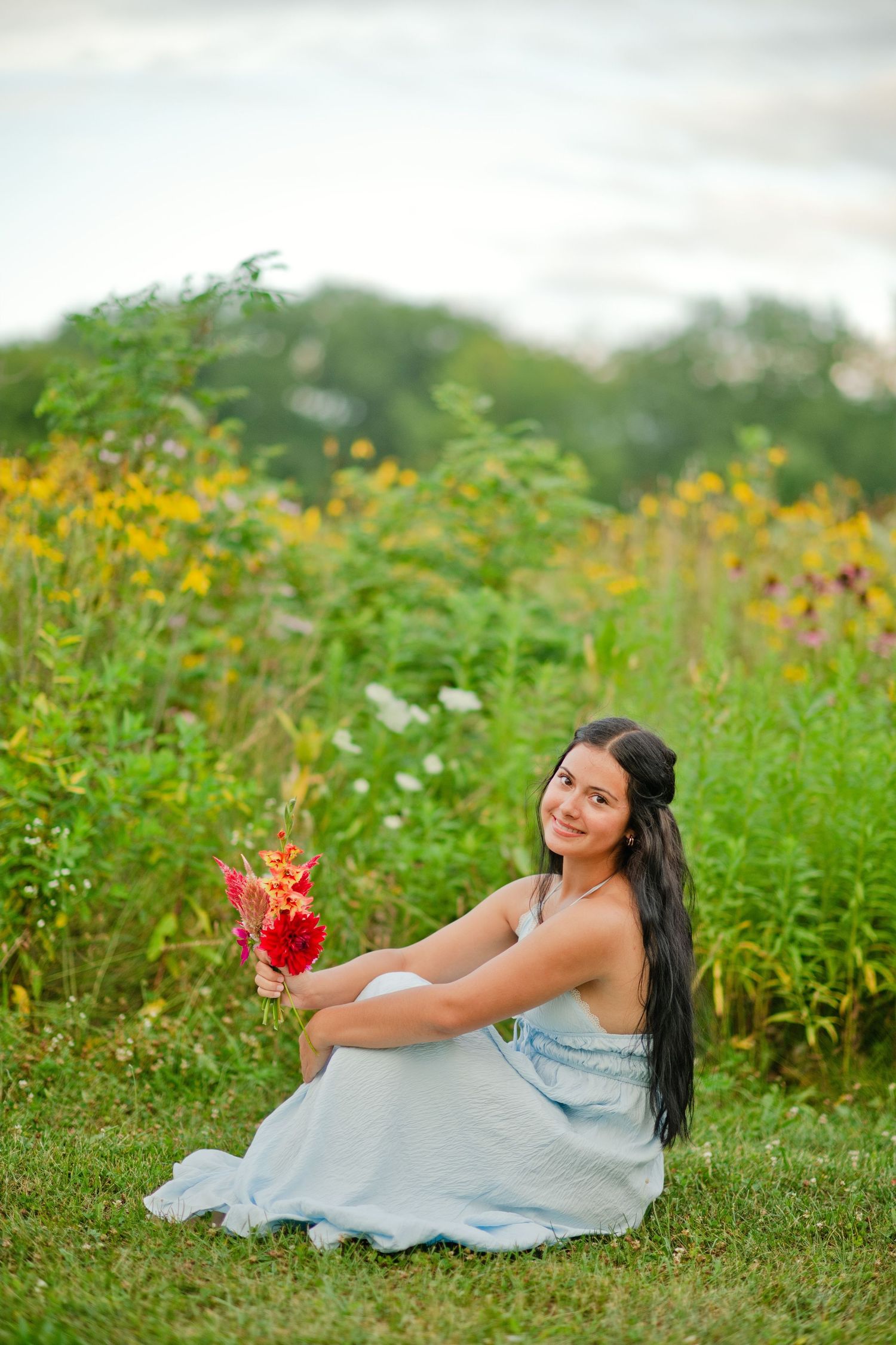 A dark-haired woman in a light blue dress sits on grass holding red flowers before a wildflower field.