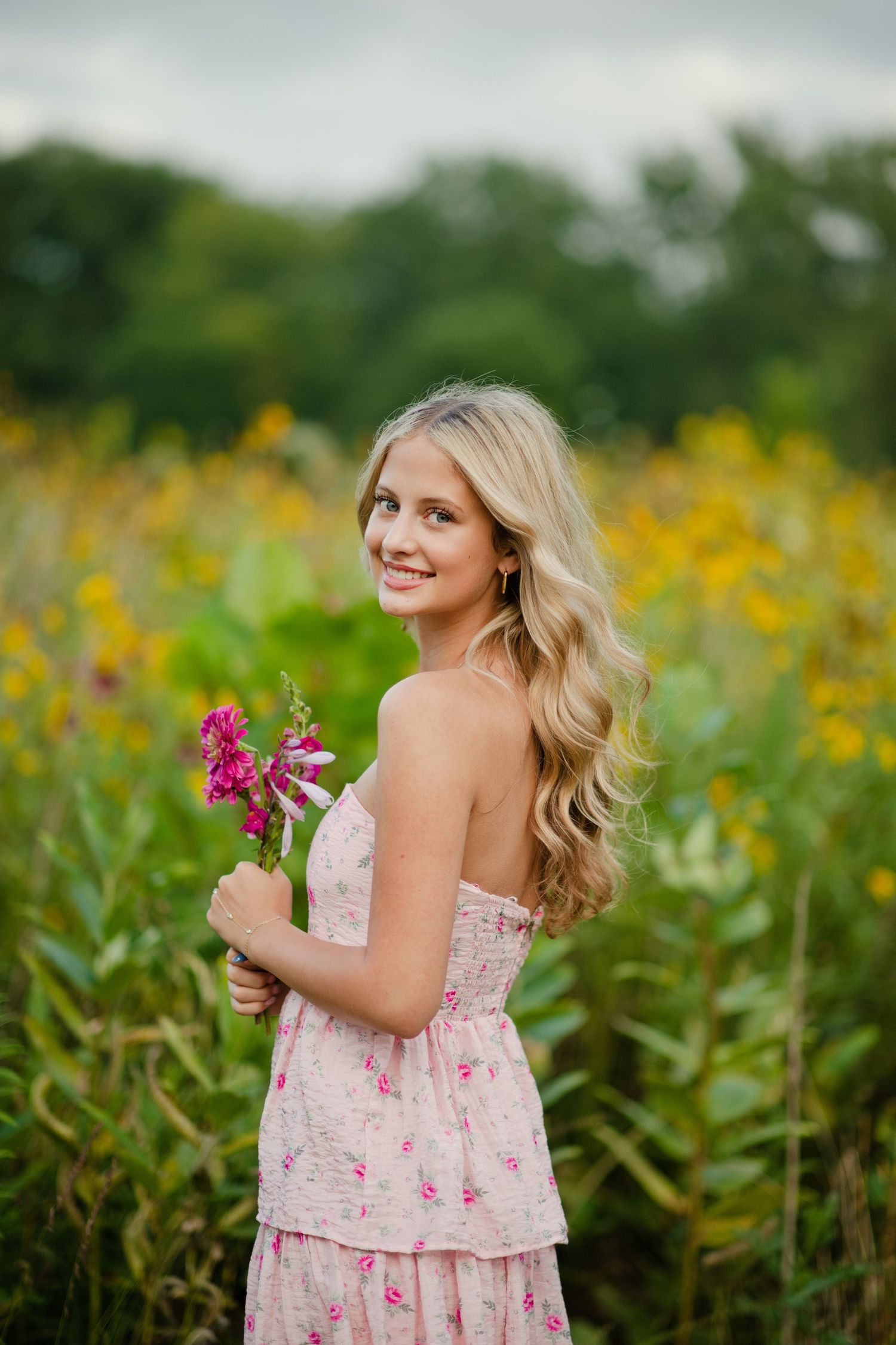 A blonde woman in a pink floral dress holds pink flowers while posing in a lush wildflower meadow.