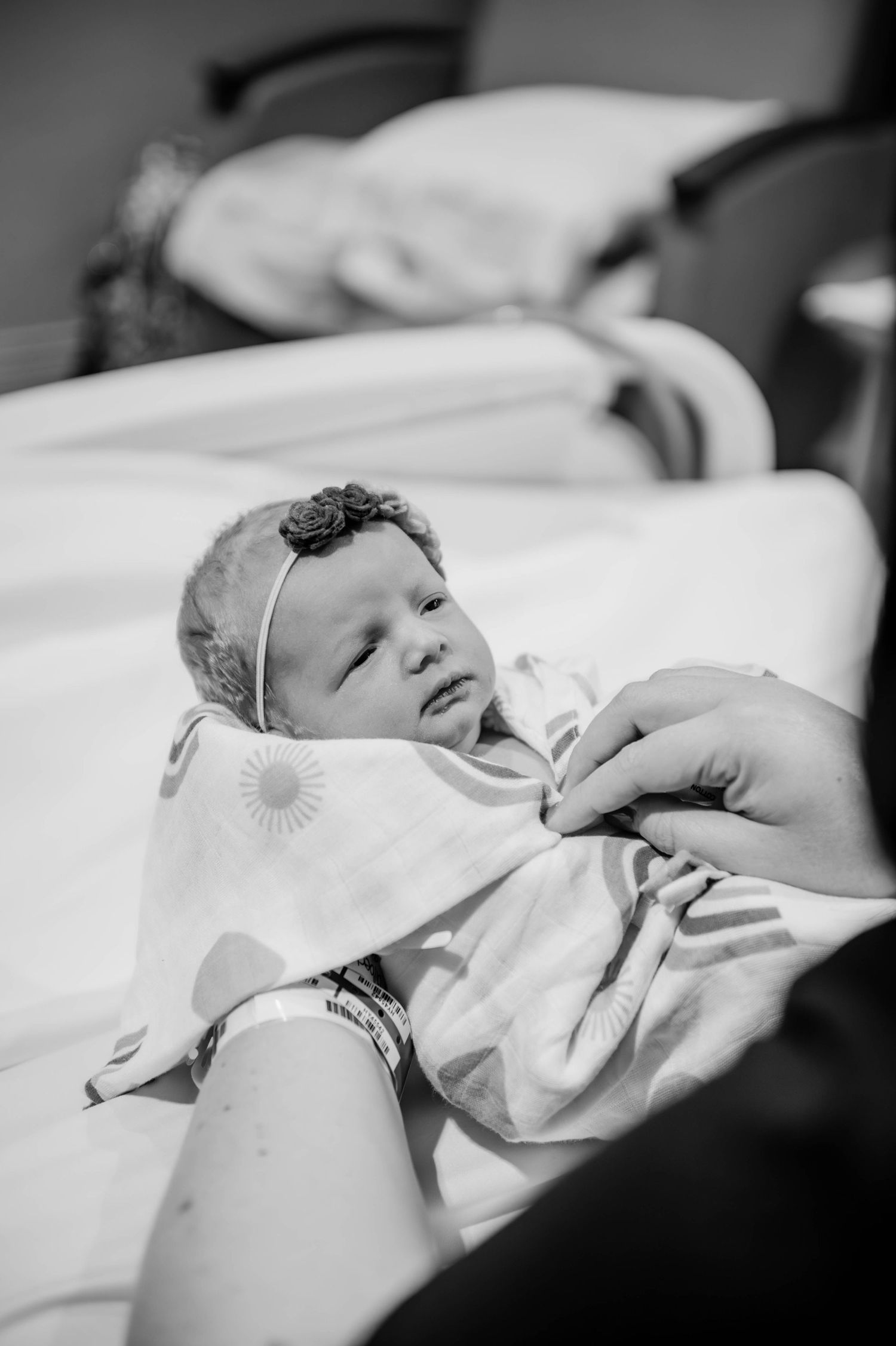 Gentle moment as a tiny newborn with floral headband rests in their hospital bassinet.