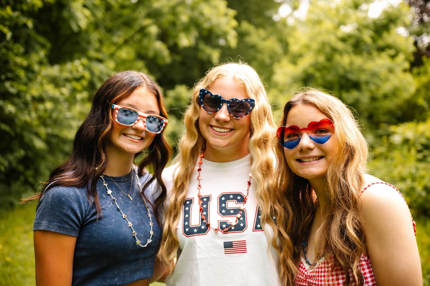 Three friends wearing patriotic themed clothing and sunglasses smile together outdoors.
