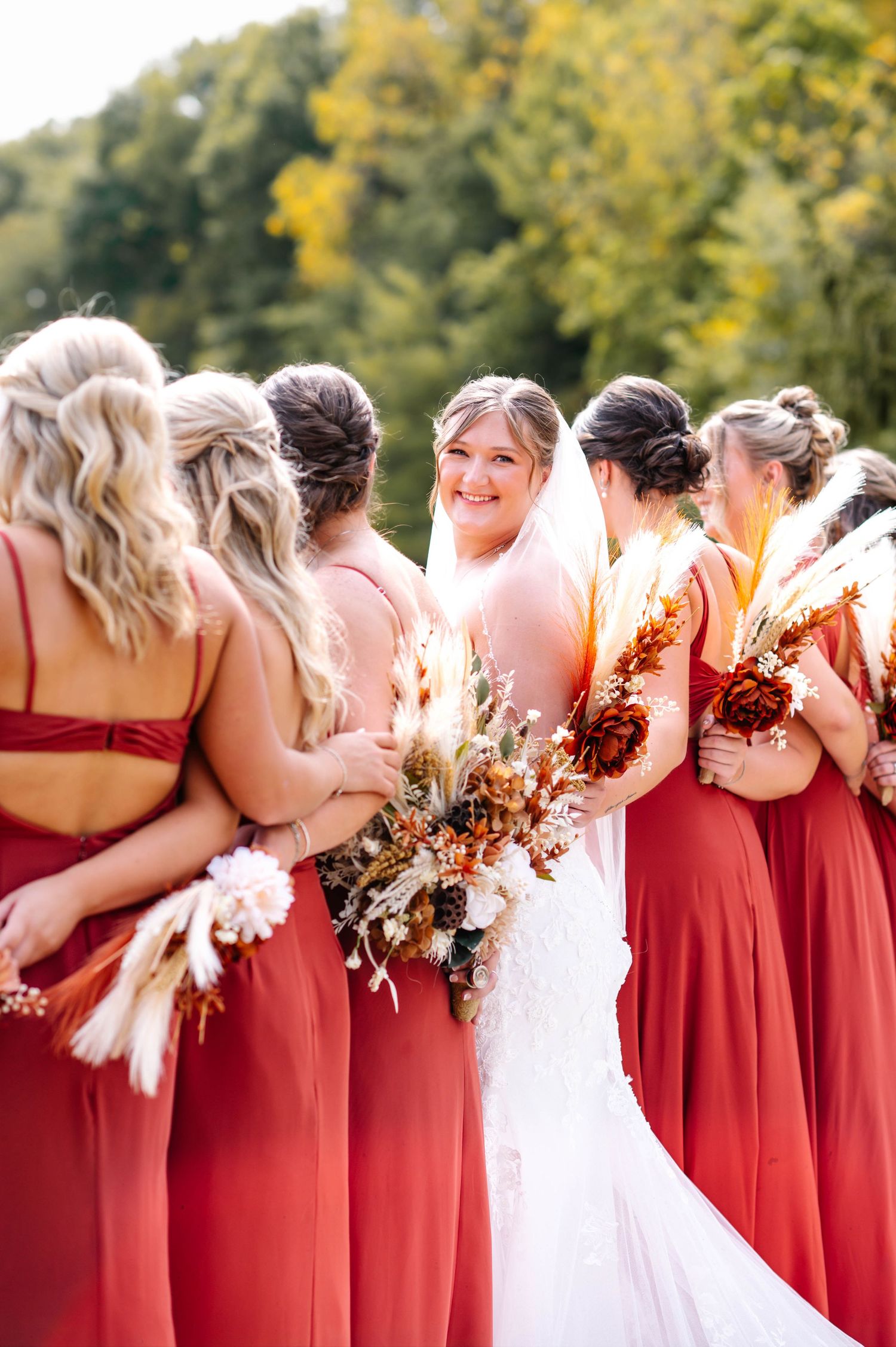 Bridal party in red dresses stands together holding bouquets at outdoor wedding.