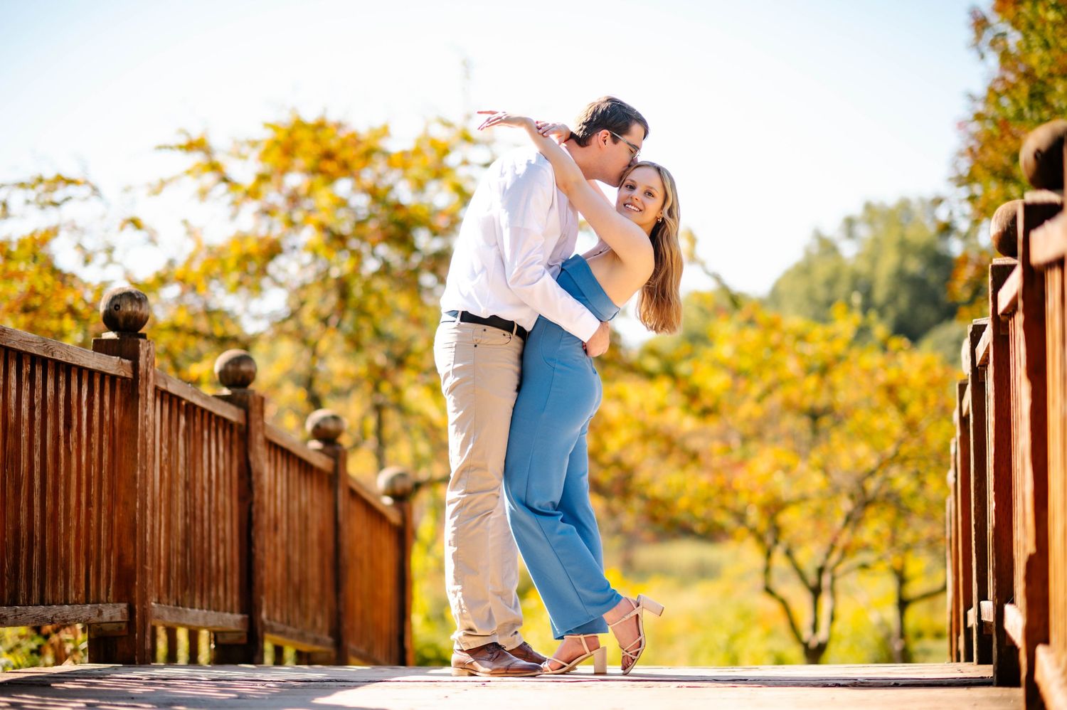 A couple embracing and kissing on a wooden bridge with autumn foliage in the background.