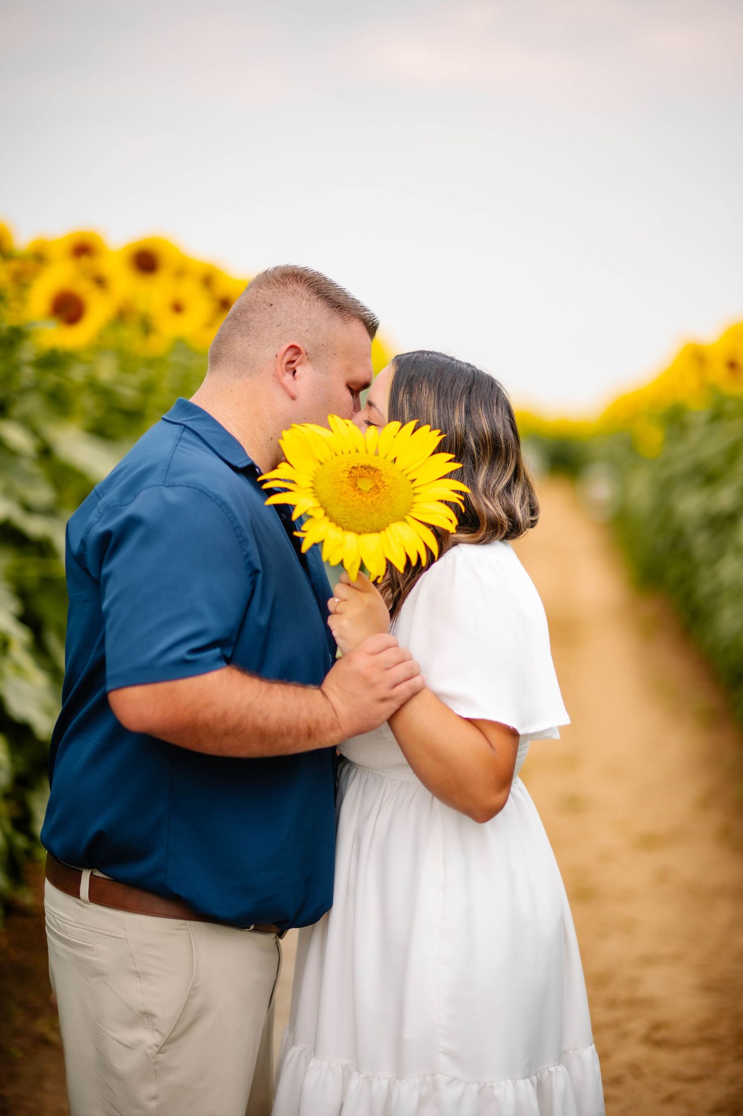 A couple sharing a romantic moment behind a large sunflower in a sunny field.