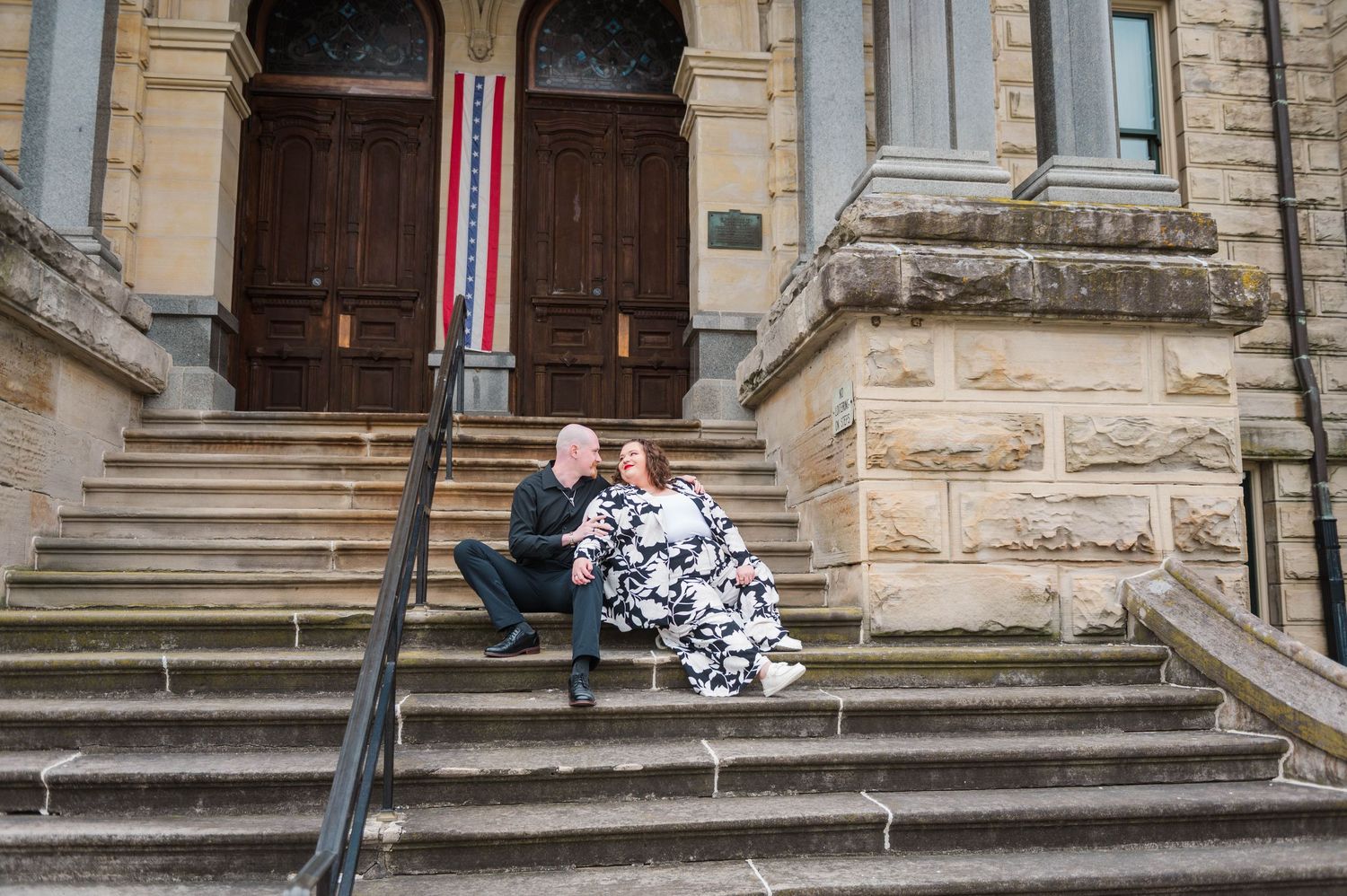 Couple in formal attire sitting on stone steps outside a historic sandstone building with ornate wooden doors.