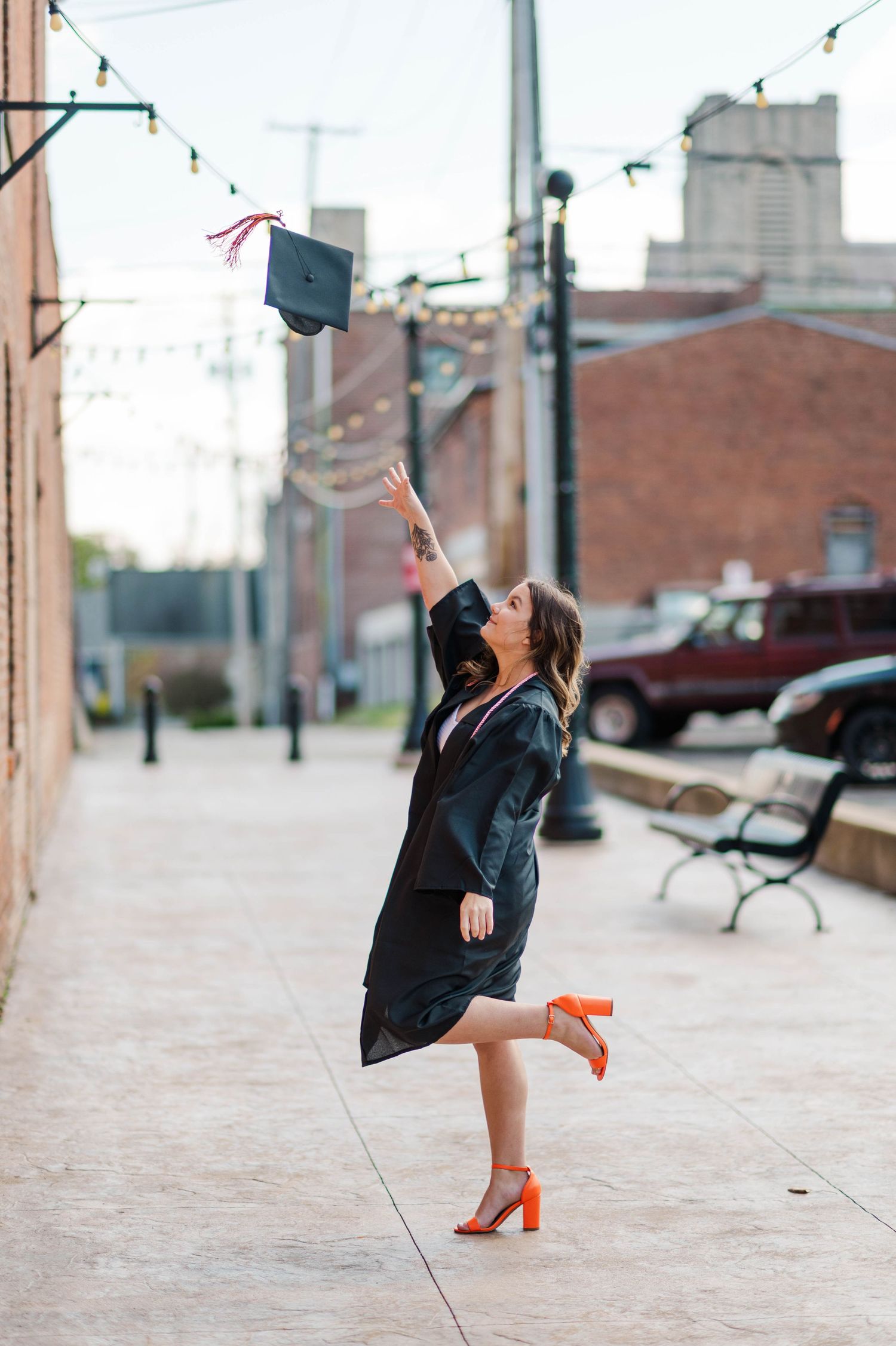 Joyful graduate tossing cap in air wearing black gown and orange heels on city sidewalk.