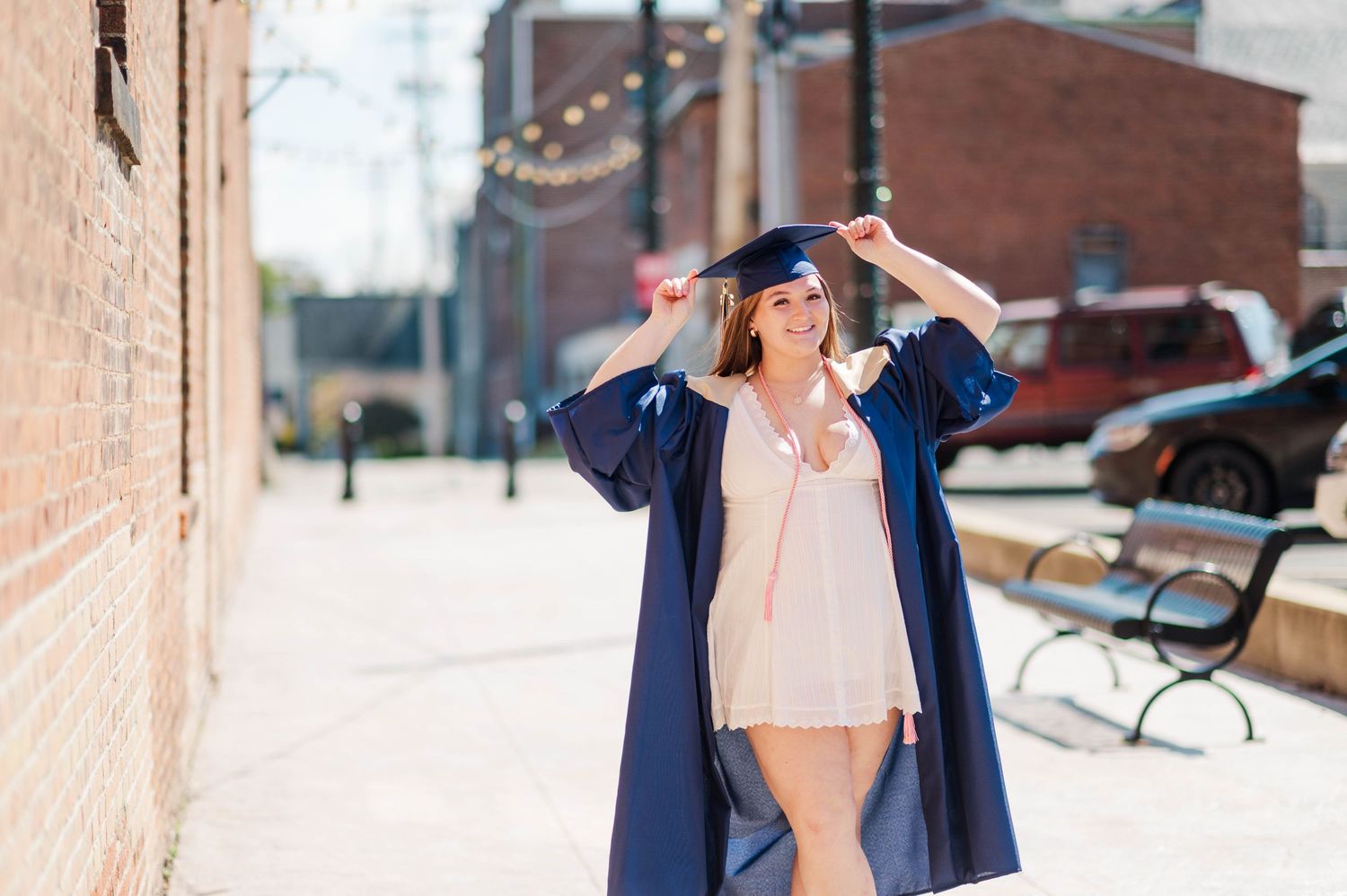 Joyful graduate in navy blue cap and gown walking outdoors adjusting her cap on sunny day.