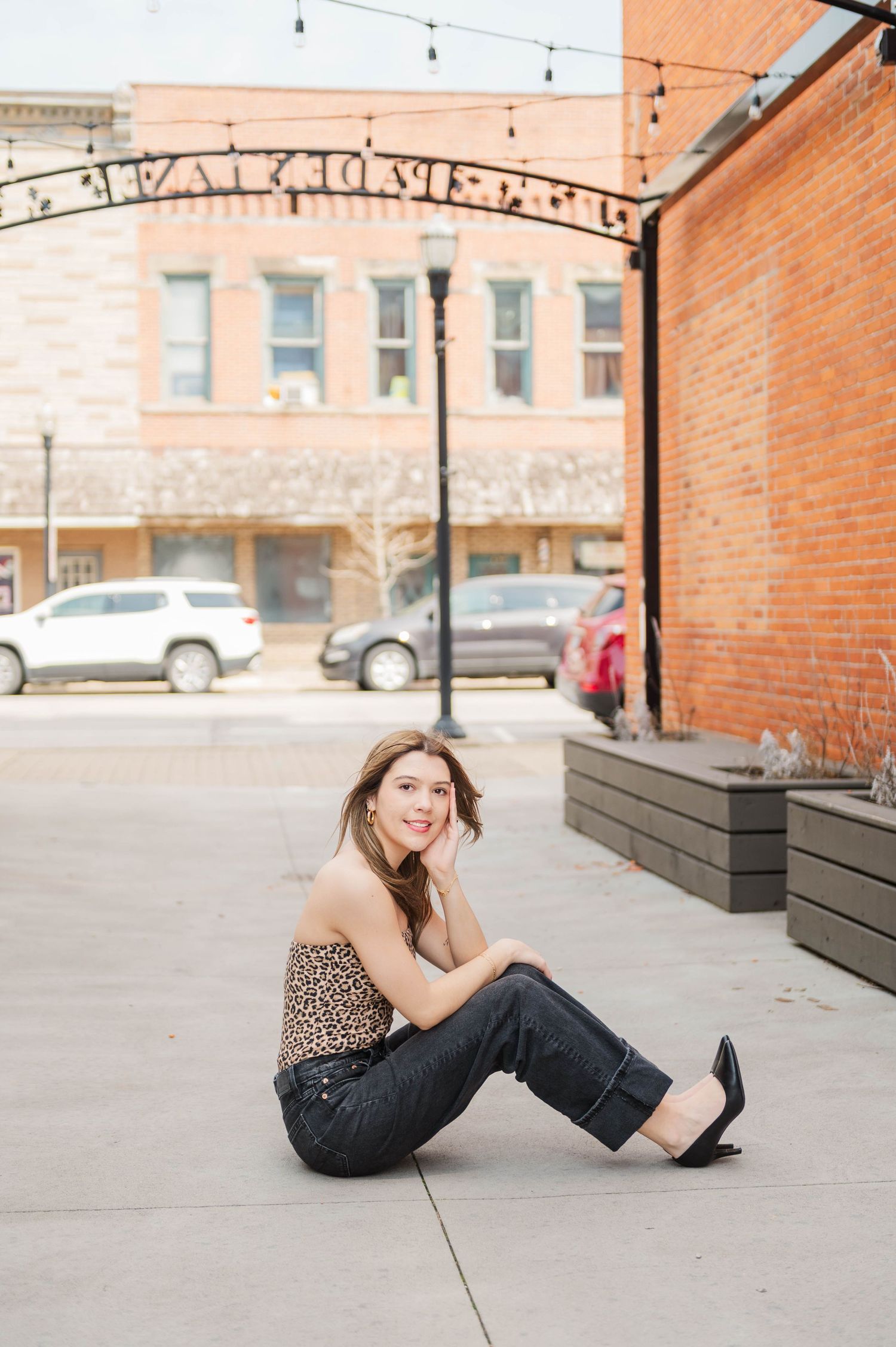 Young woman in leopard print top and dark jeans sitting on a sidewalk under a decorative arch.