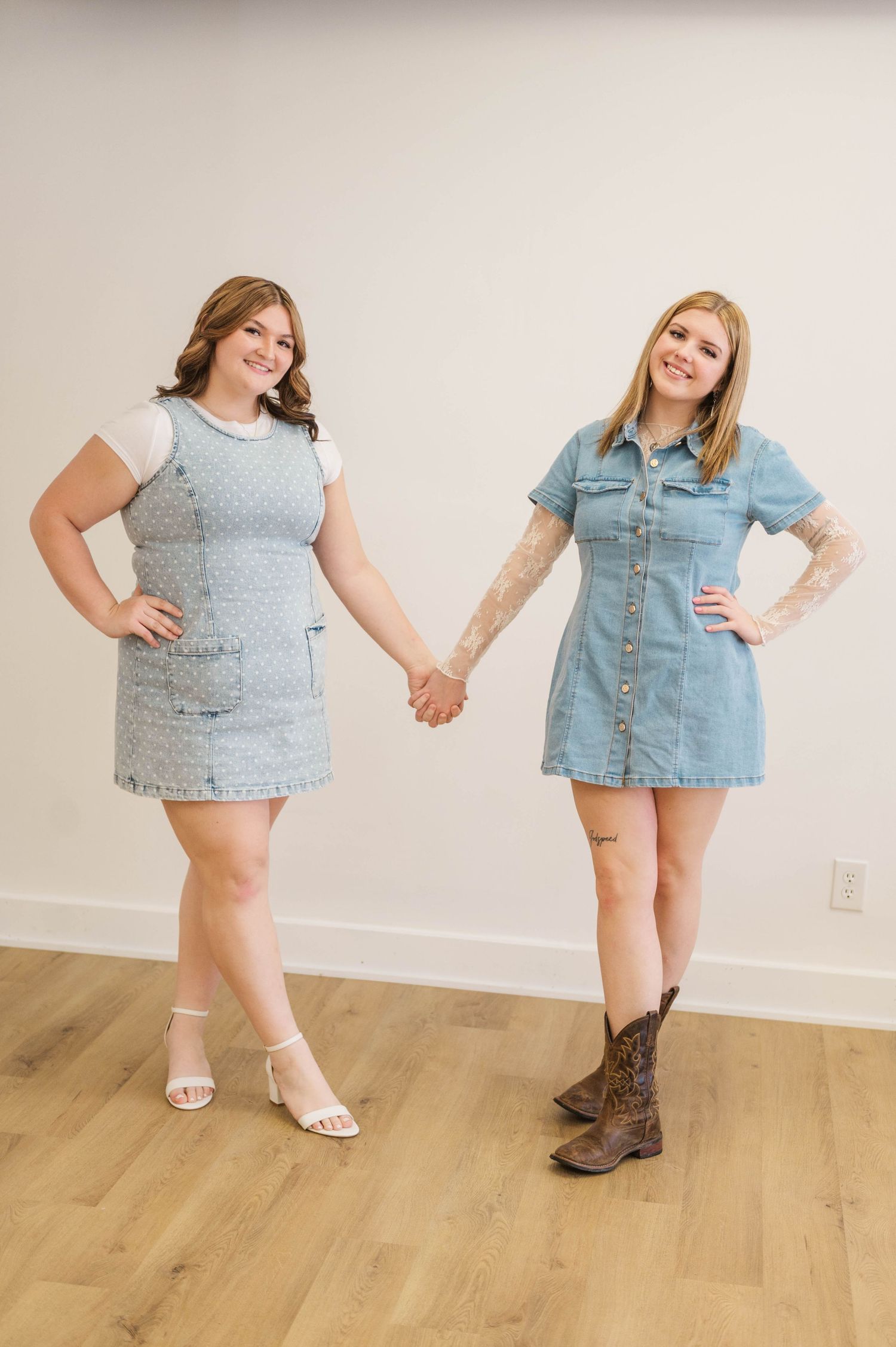 Two women in denim dresses holding hands and smiling in a bright minimalist studio.