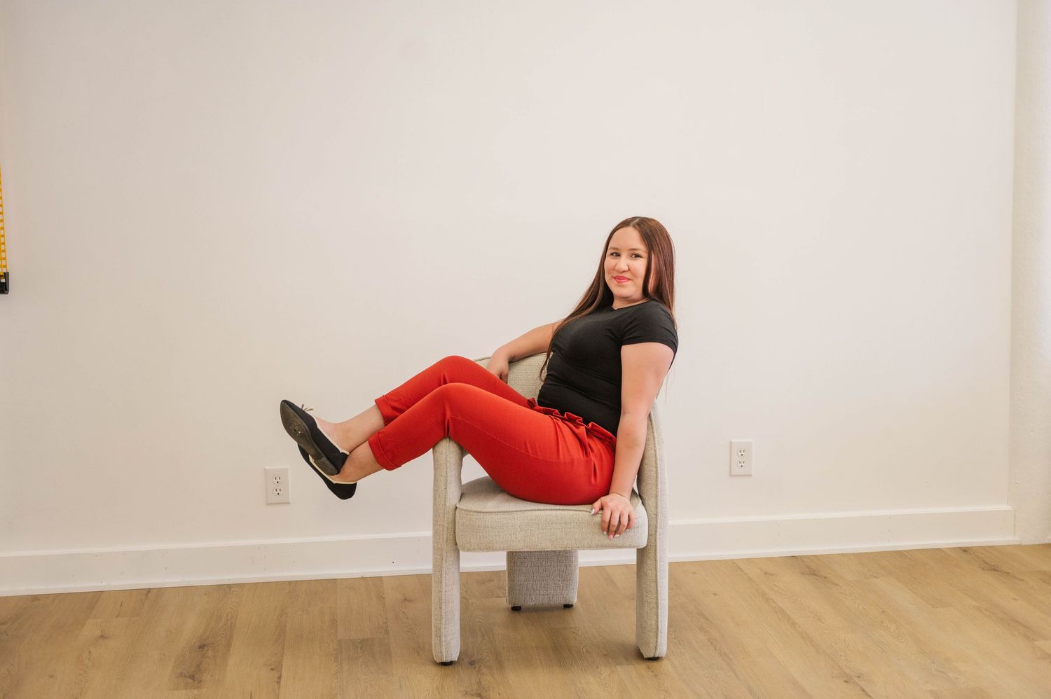 Brunette woman in black tee and red pants sitting casually in a white modern chair.