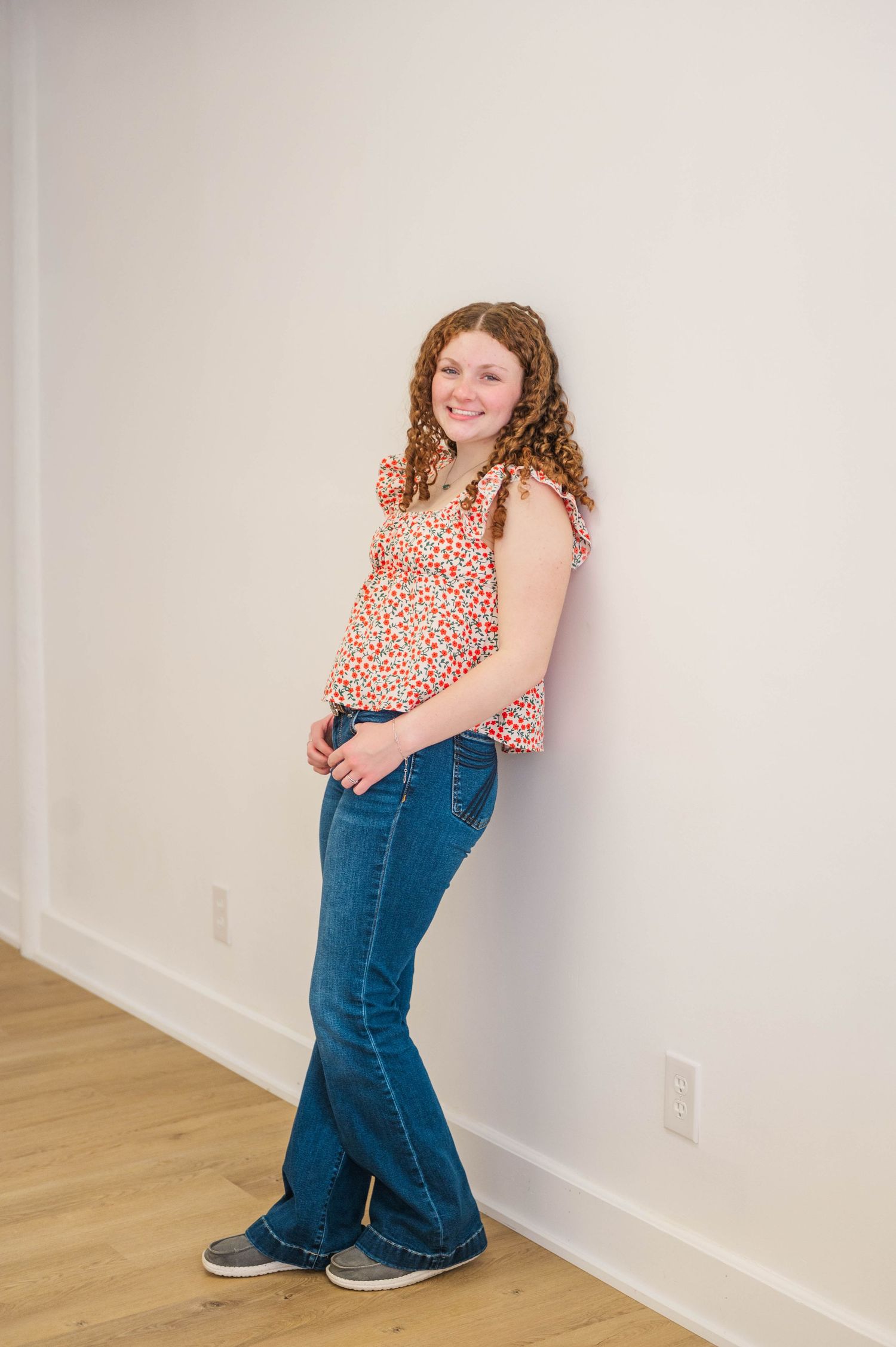 Curly redhead girl leans against white wall in floral top and flared jeans, smiling warmly.