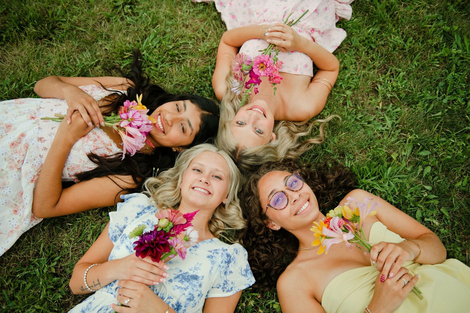 Four young women in floral dresses lying in a circle on grass holding colorful flowers.