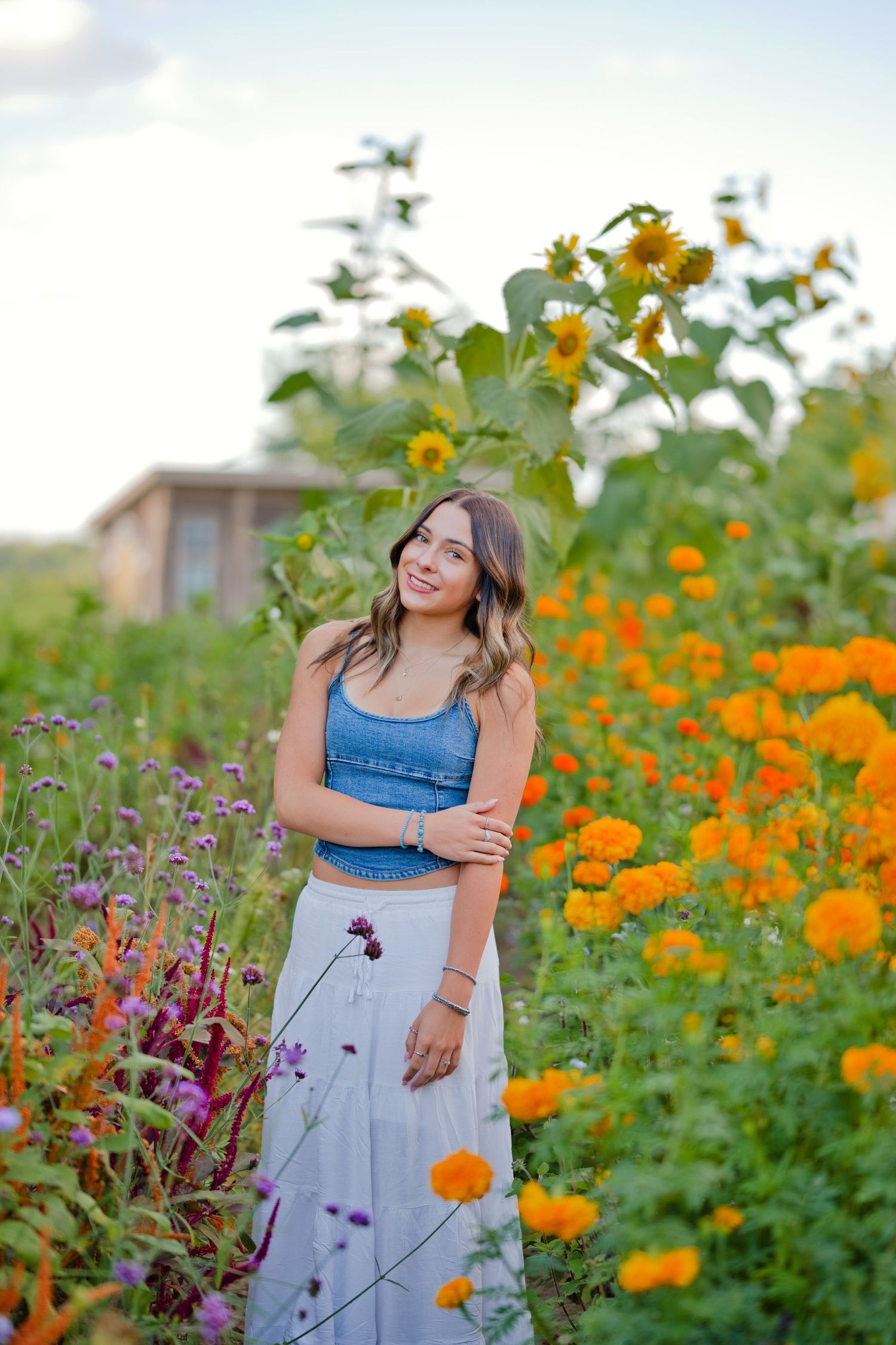 Young woman in denim crop top and white skirt smiling among vibrant orange marigolds and sunflowers in a garden.