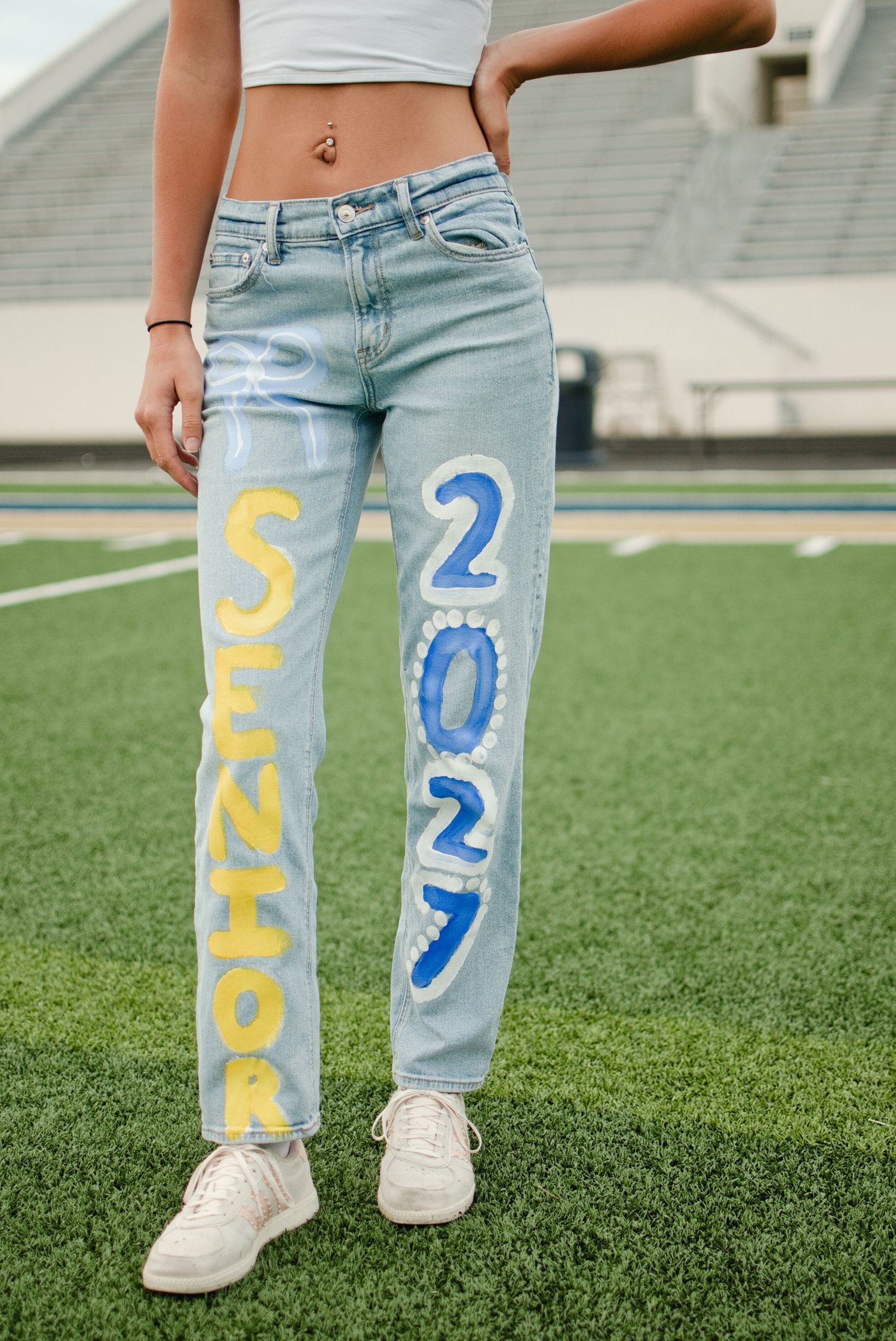Close-up of light wash jeans painted with yellow SENIOR and blue 2027 on a football field.