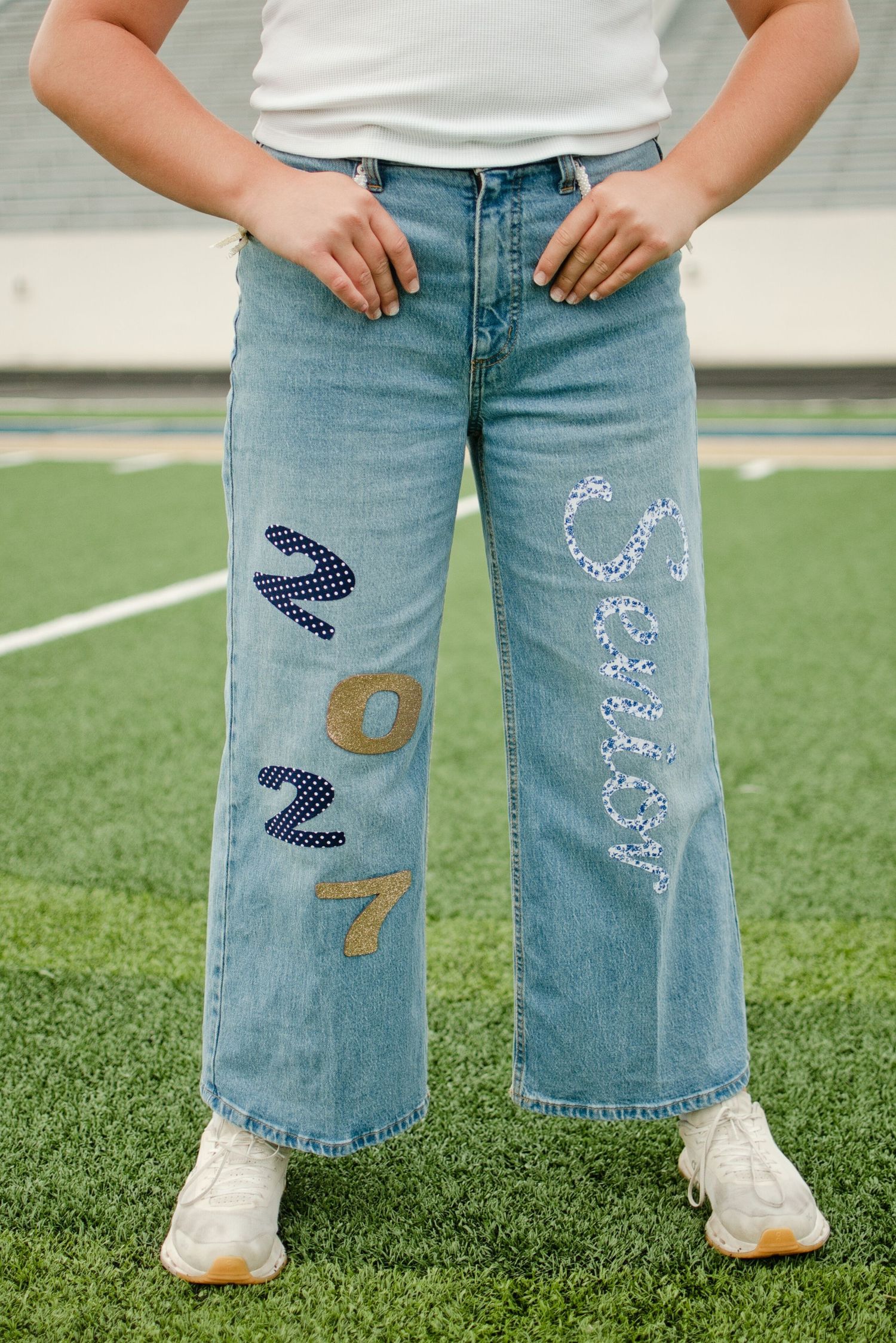 Close-up of wide-leg jeans with embroidered 2027 and name patches on a green football field.