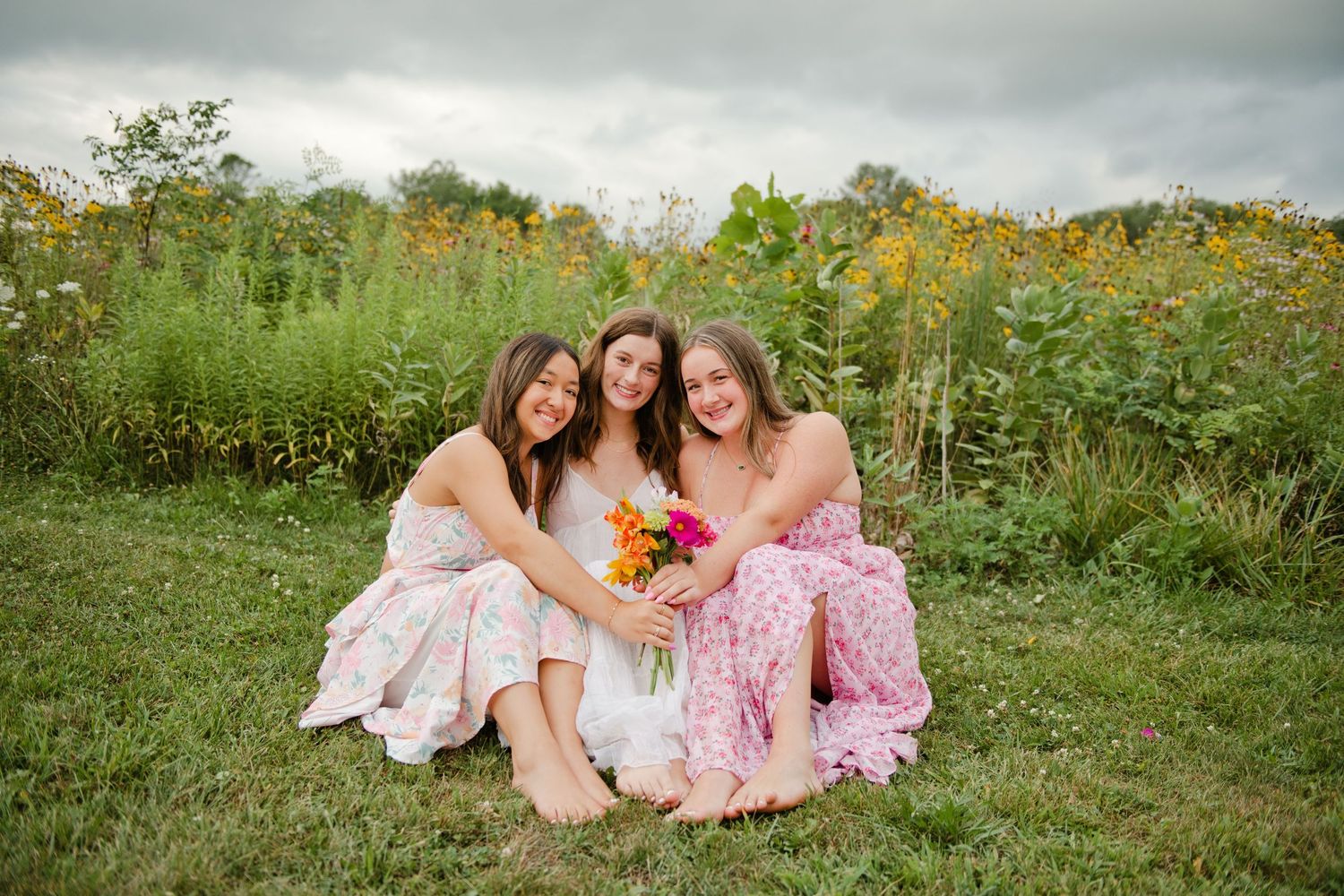 Three smiling women in floral pink and white dresses sitting together in a wildflower meadow.