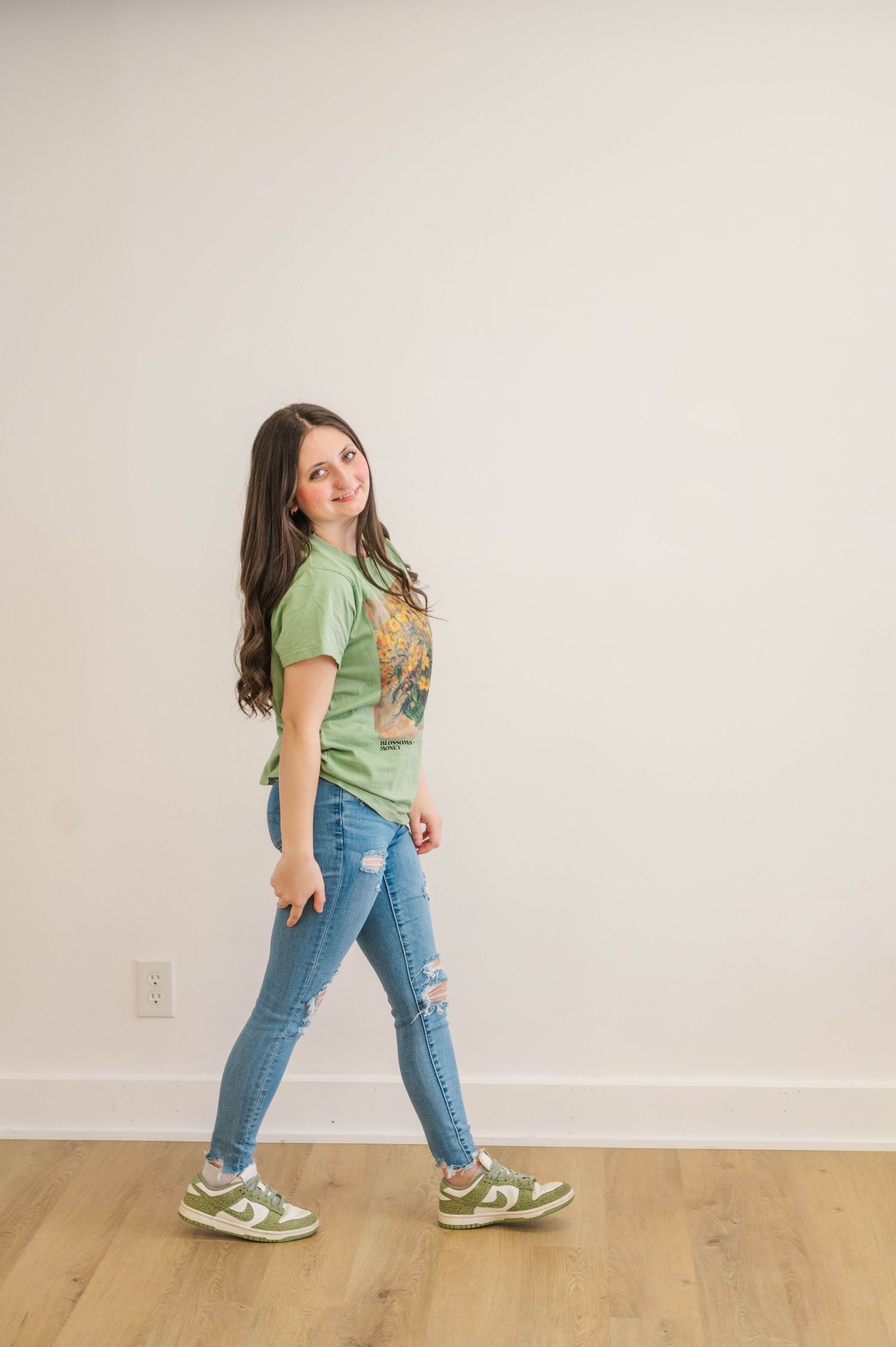 Brunette woman in green graphic tee and ripped jeans walking on hardwood floor against white wall.