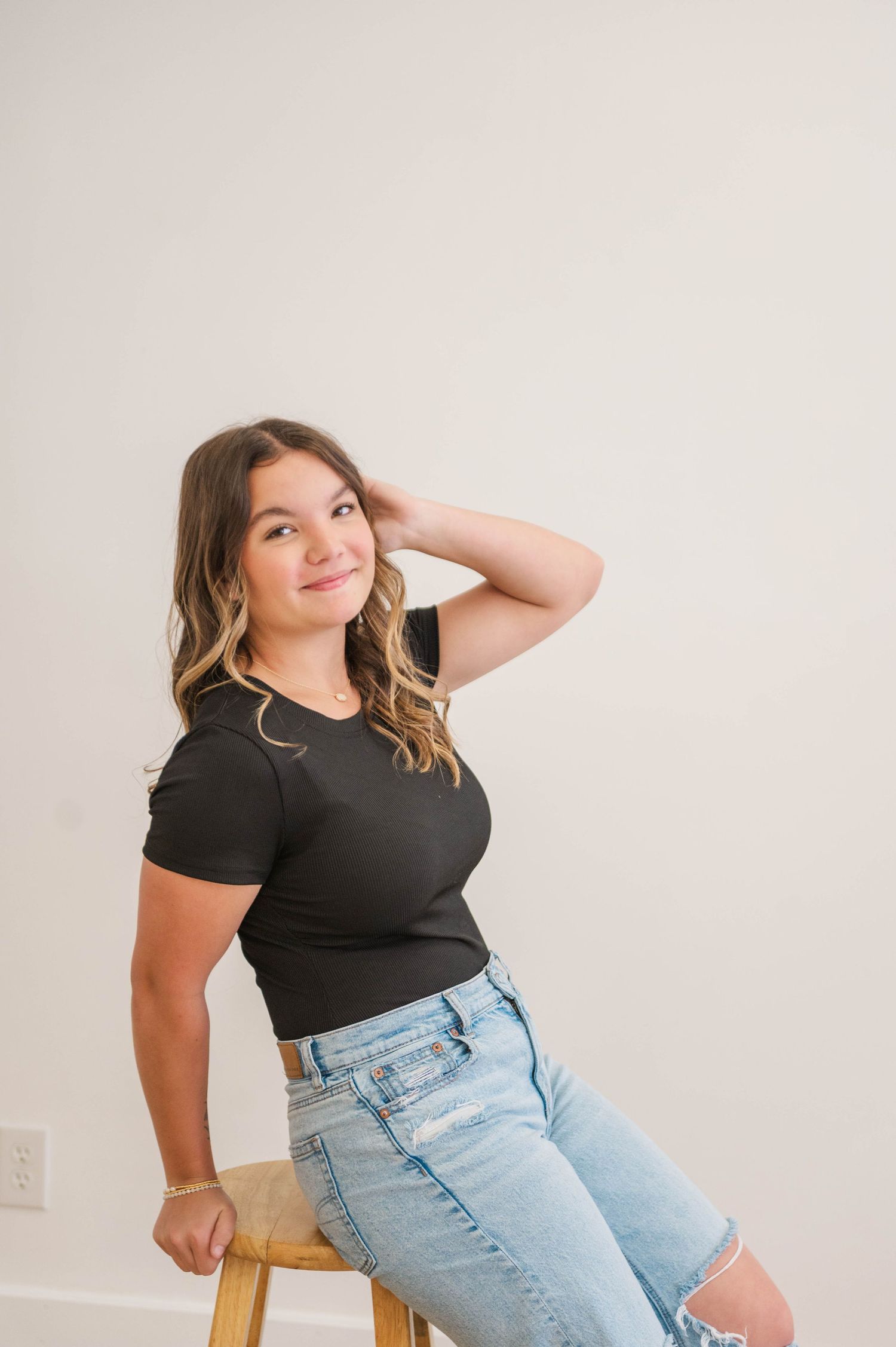 Young woman in black tee and light wash jeans sitting on stool, smiling over shoulder.