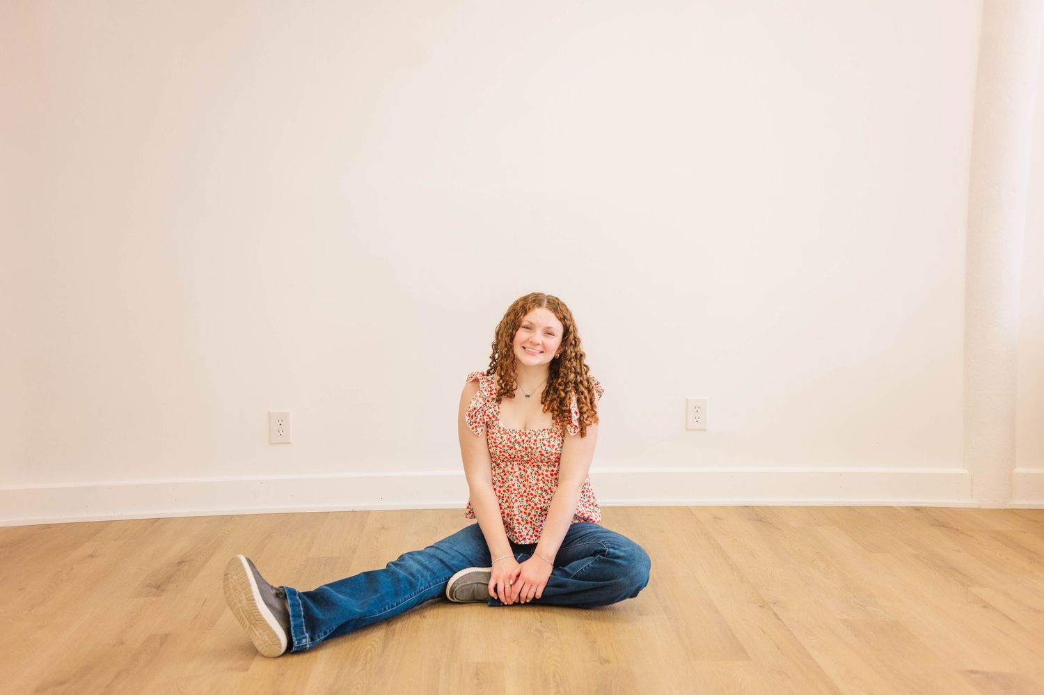 Young girl with curly red hair sitting cross-legged on hardwood floor, wearing jeans and floral top, smiling.