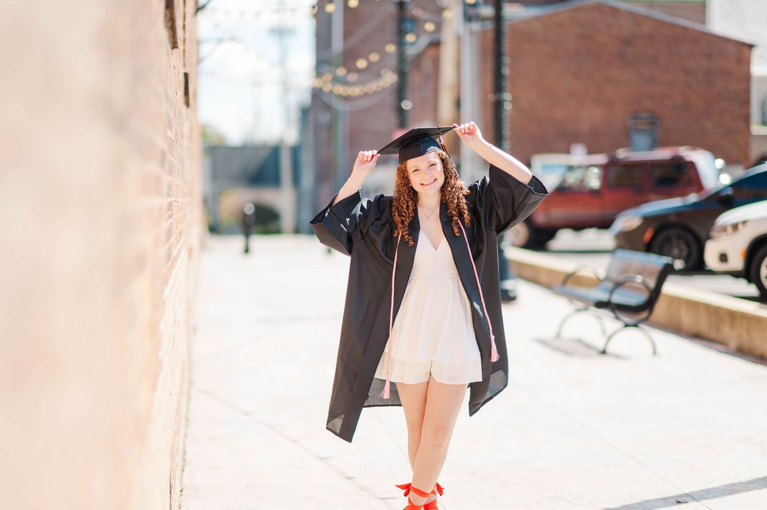 Graduate adjusting black cap and gown on sunny brick-lined city street.