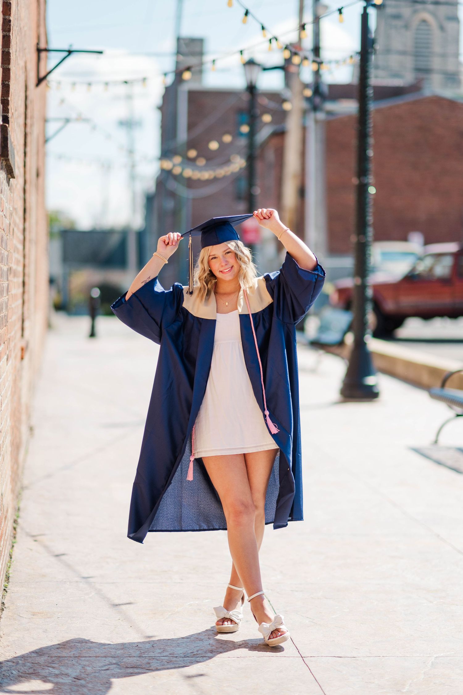 Smiling blonde graduate in navy cap and gown walking on urban sidewalk with string lights overhead.