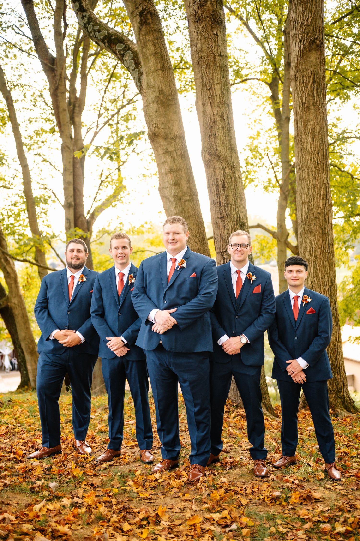 Groomsmen in navy suits pose playfully on fallen autumn leaves.