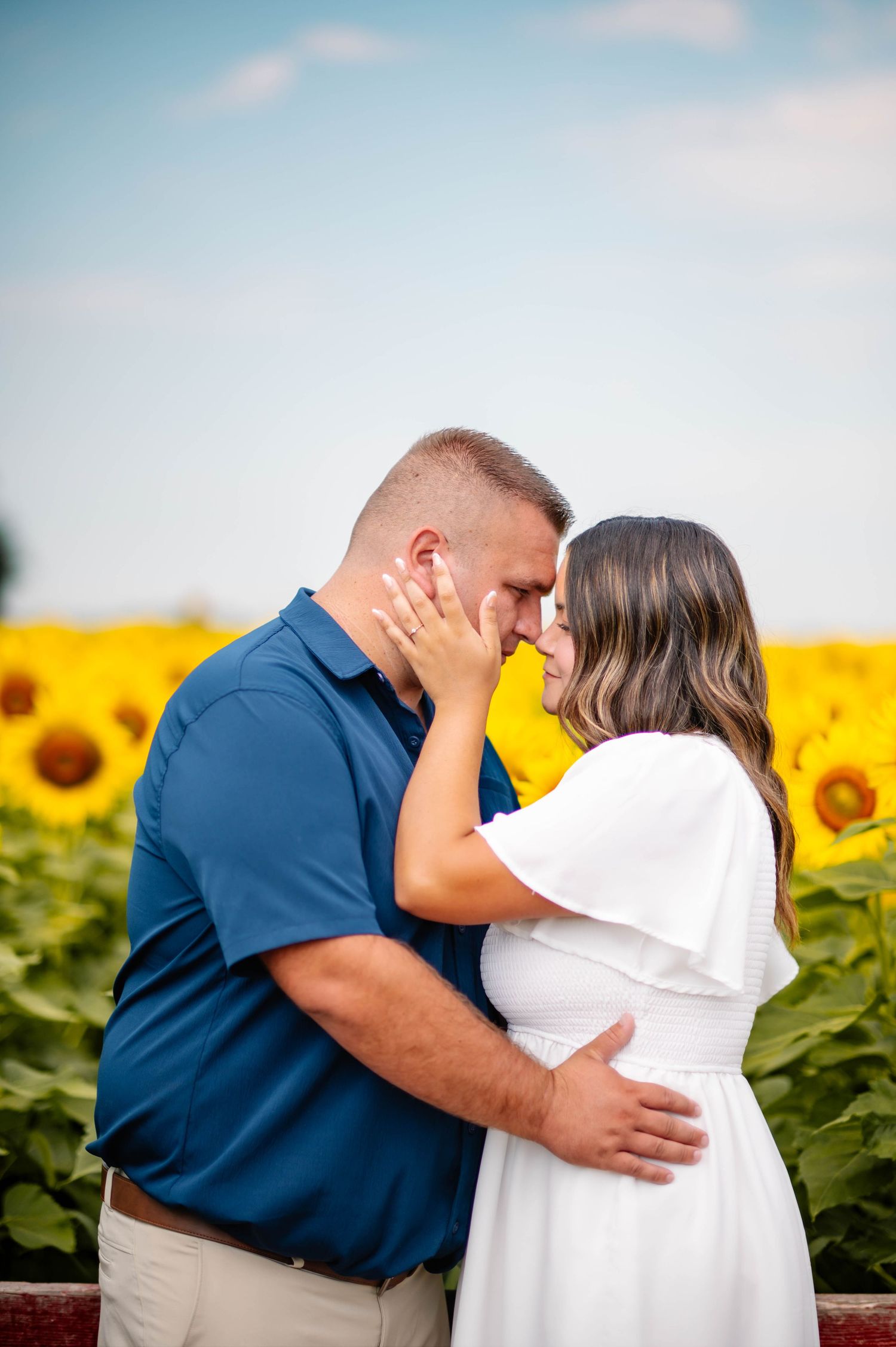 A couple embraces in a sunflower field against a bright blue sky.
