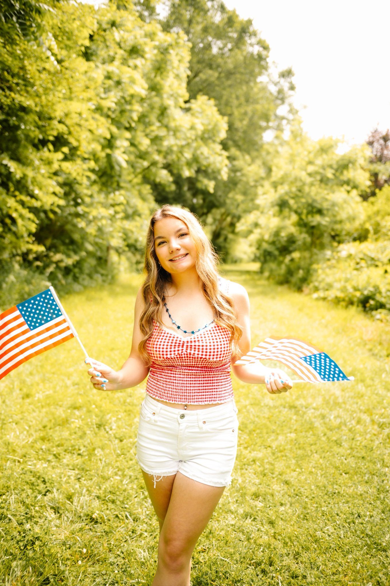 Woman in gingham top waves American flags while posing in sunny outdoor setting.