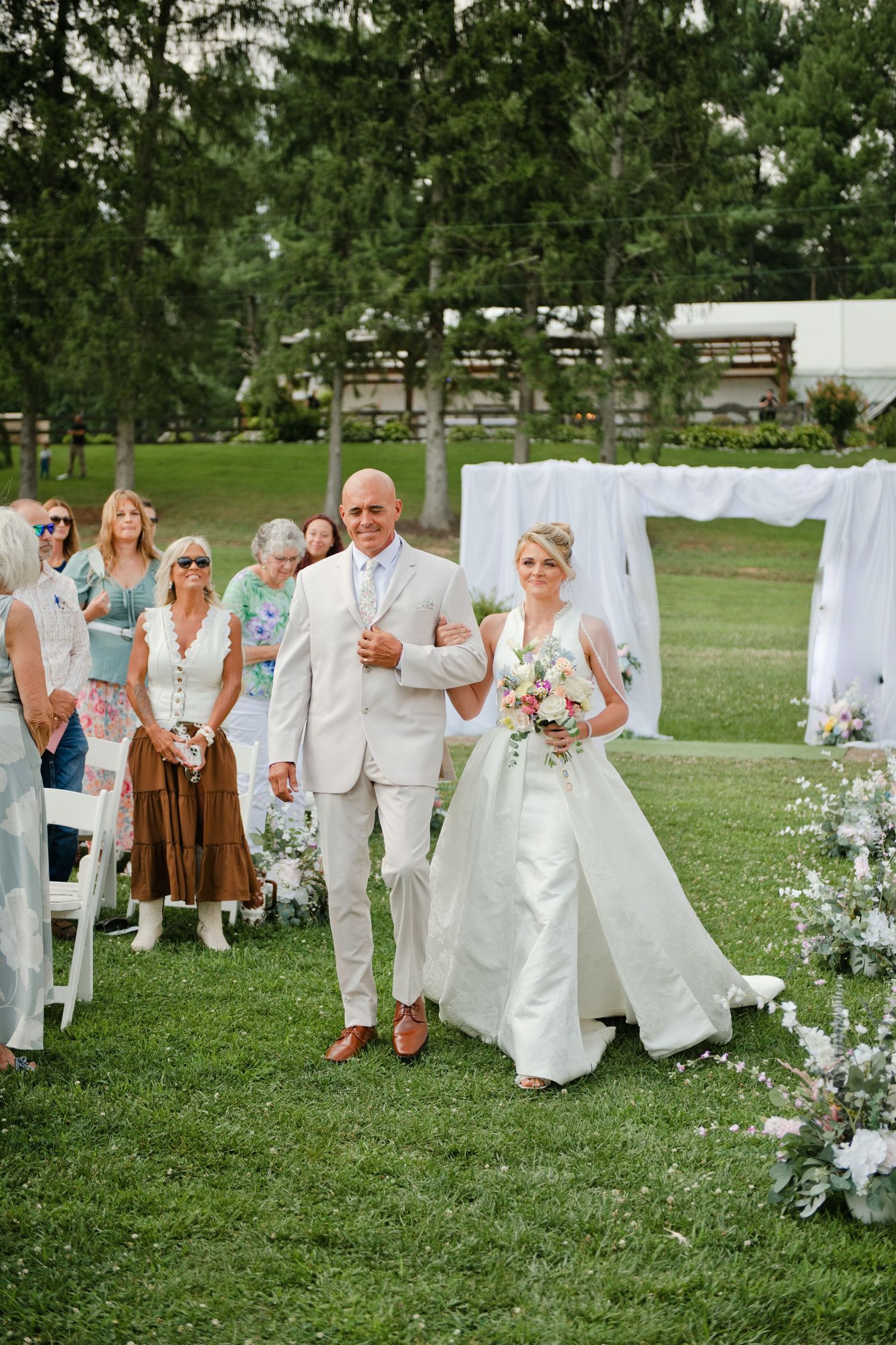 Bride and groom walk down outdoor wedding aisle, surrounded by guests, white draping, and floral arrangements on green lawn.