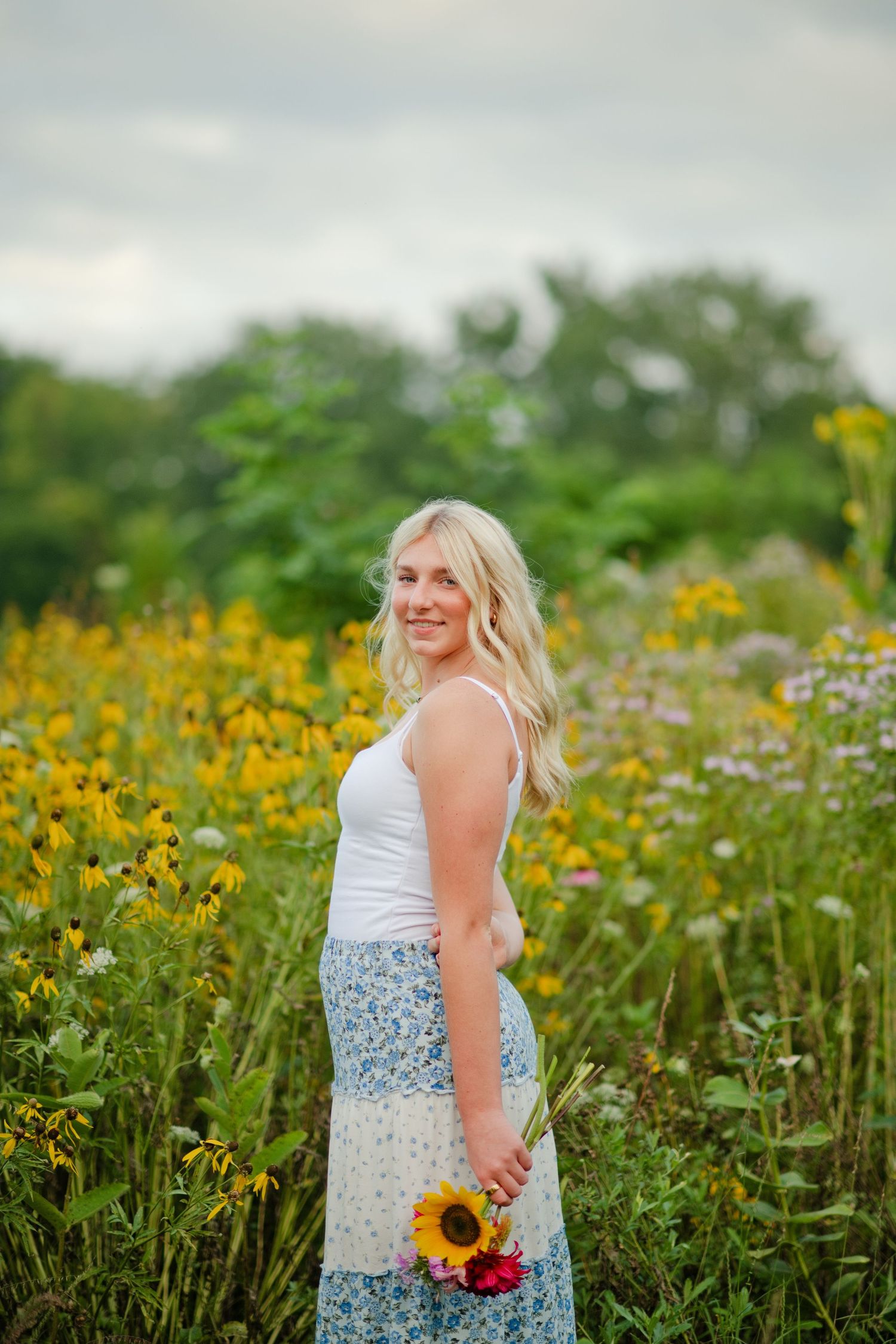Smiling blonde woman in a blue floral skirt holding a sunflower bouquet in a vibrant wildflower meadow.