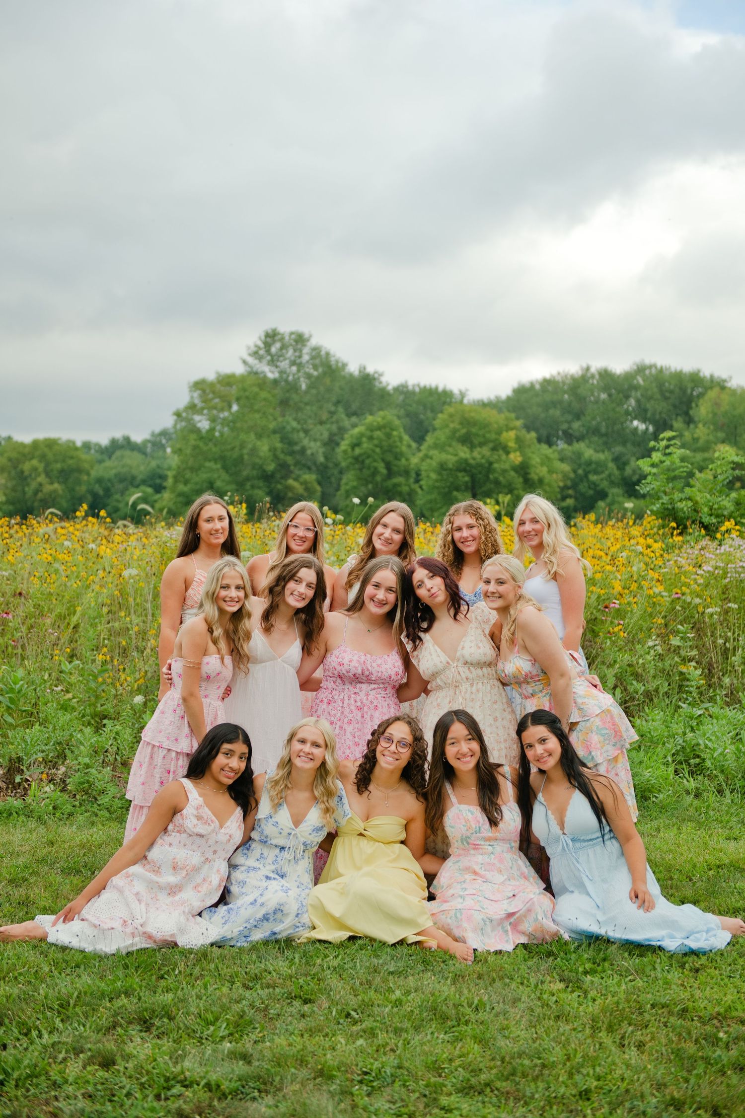 Group of young women in colorful floral dresses posing together in a sunny field with wildflowers and trees.