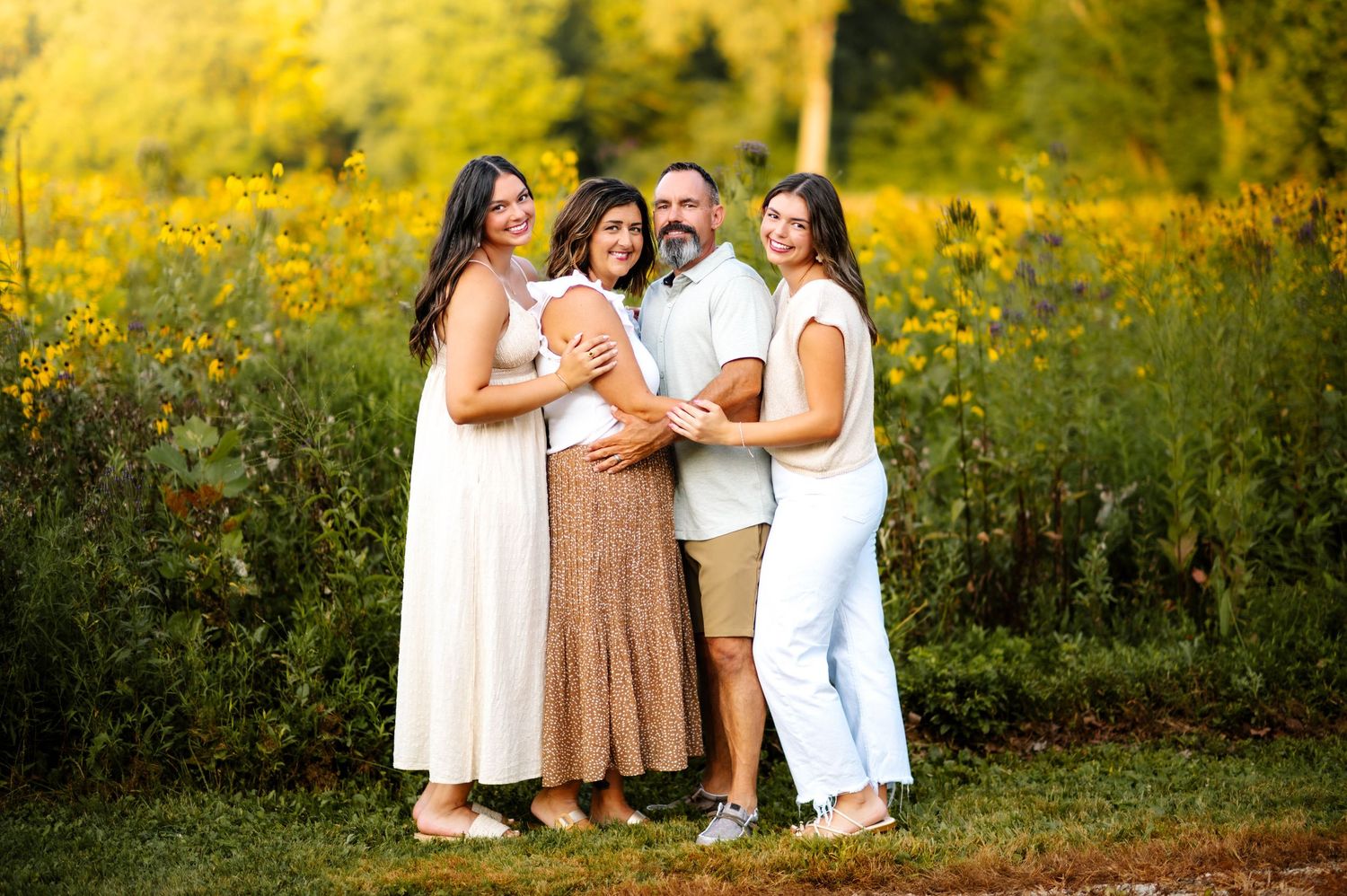 A group poses together in a golden field at sunset wearing coordinated white and neutral toned outfits.