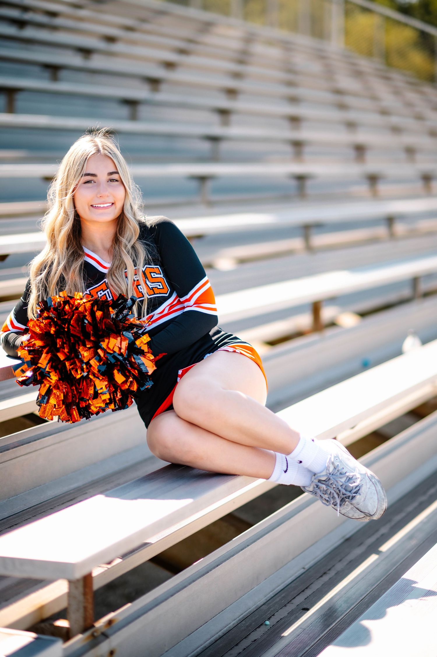 Cheerleader poses on metal bleachers while holding orange and black pom poms.