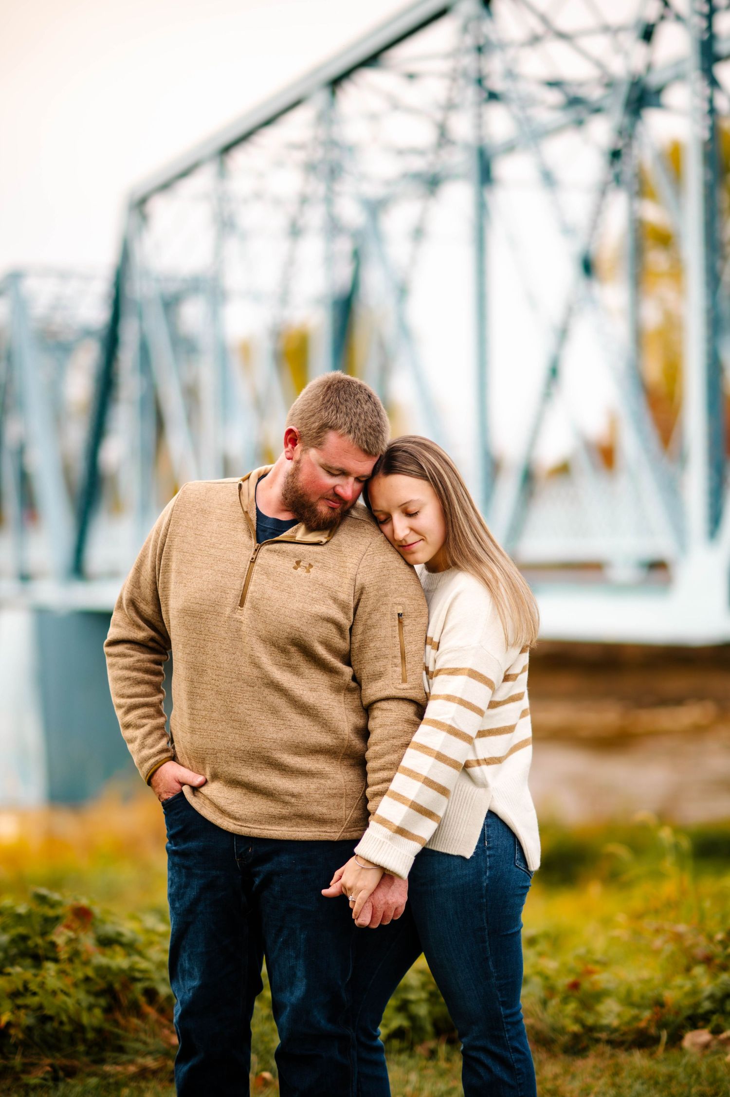 Couple in casual sweaters share tender moment outdoors with industrial bridge backdrop.