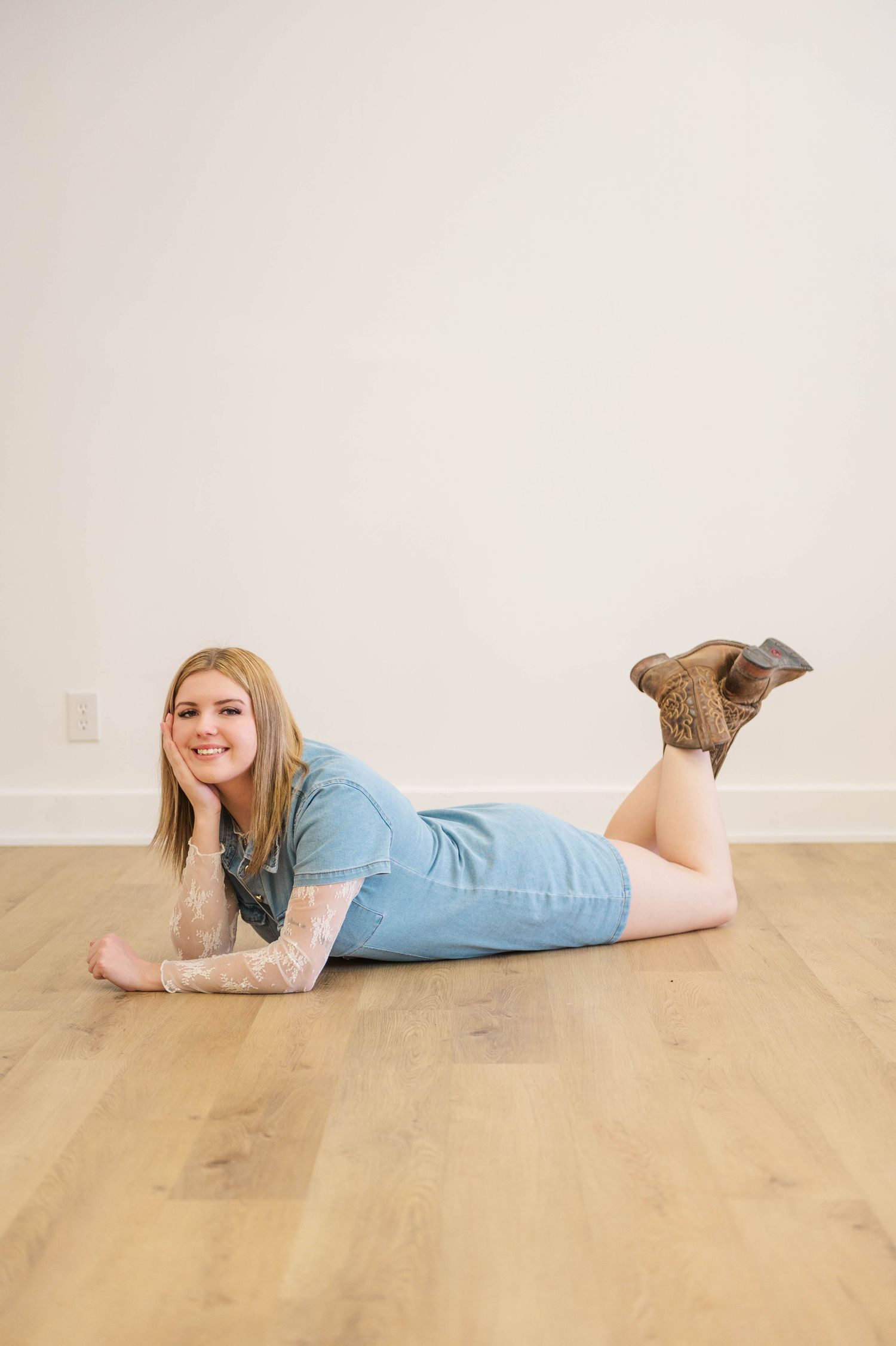 Smiling blonde woman in denim dress and cowboy boots lying on hardwood floor in bright studio.