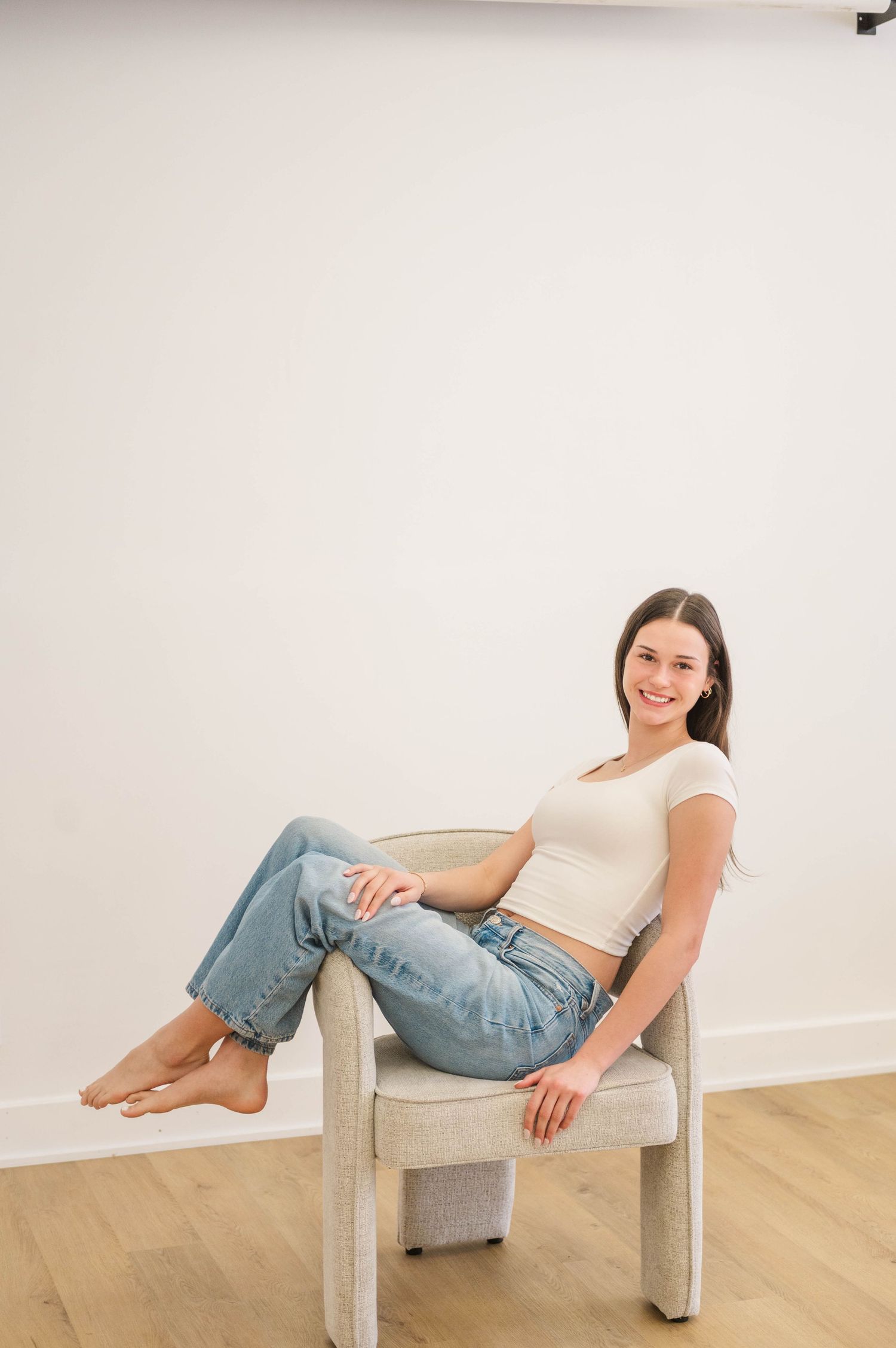 Young woman in cream top and jeans sitting barefoot in beige chair, smiling naturally.