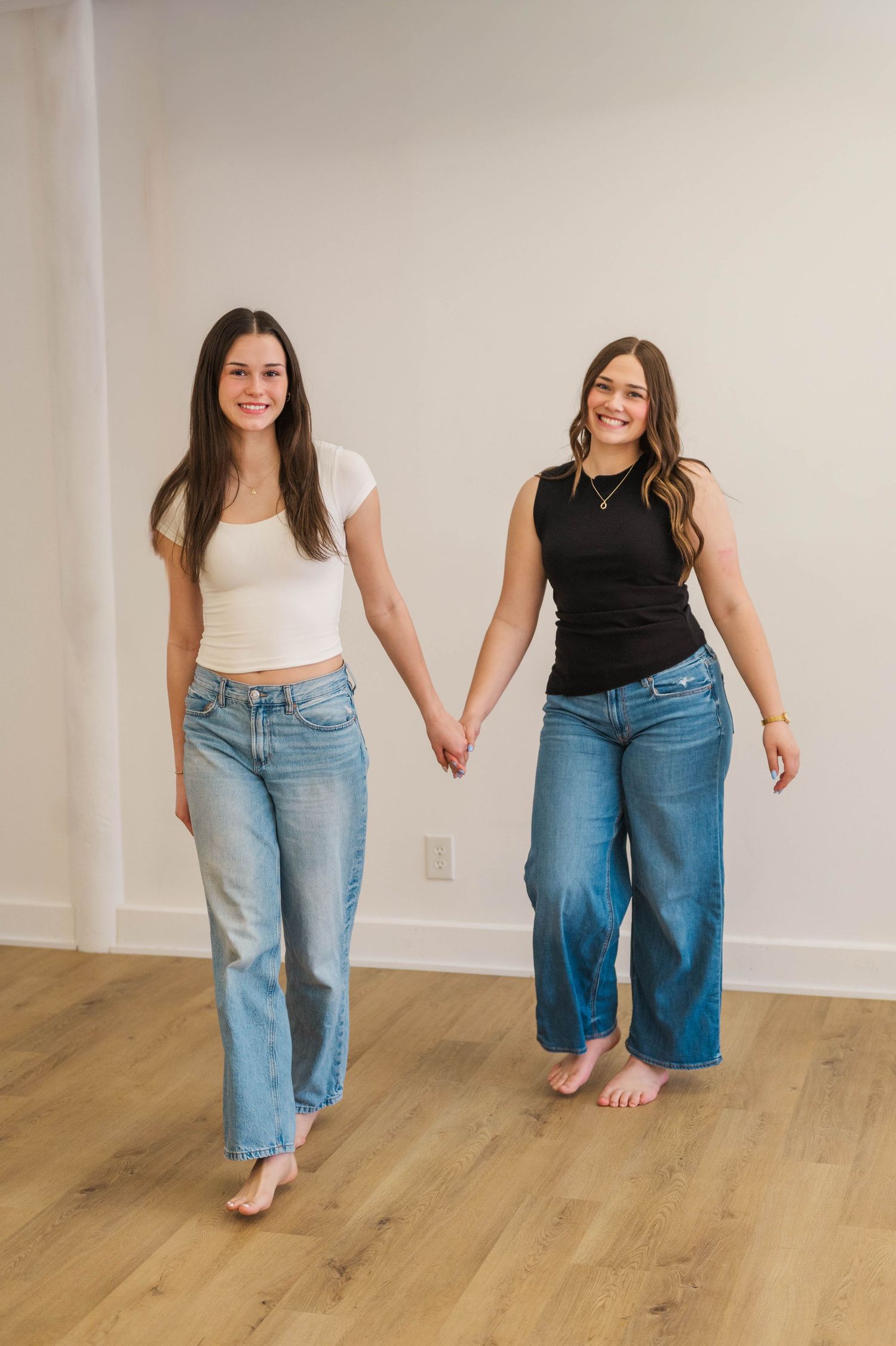 Two smiling women holding hands walking barefoot in jeans, one in white crop top and one in black tank top.
