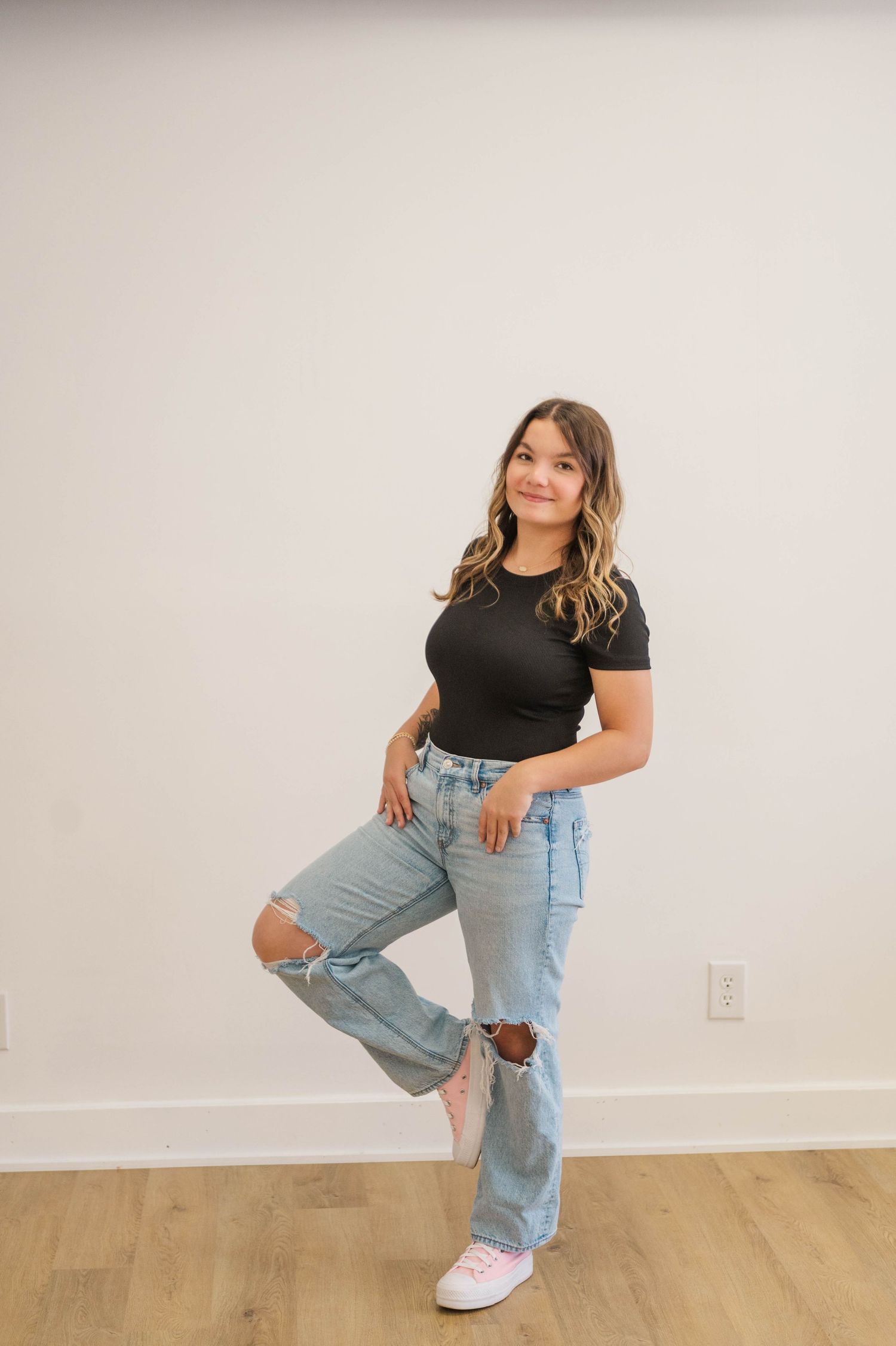 Woman in black crop tee and ripped light wash jeans standing with one knee raised.
