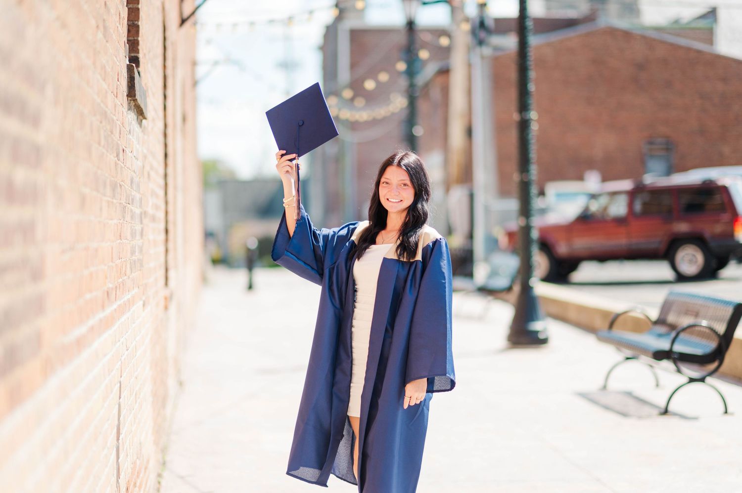 Smiling female graduate in navy blue gown raises mortarboard cap on sunny downtown brick alley.
